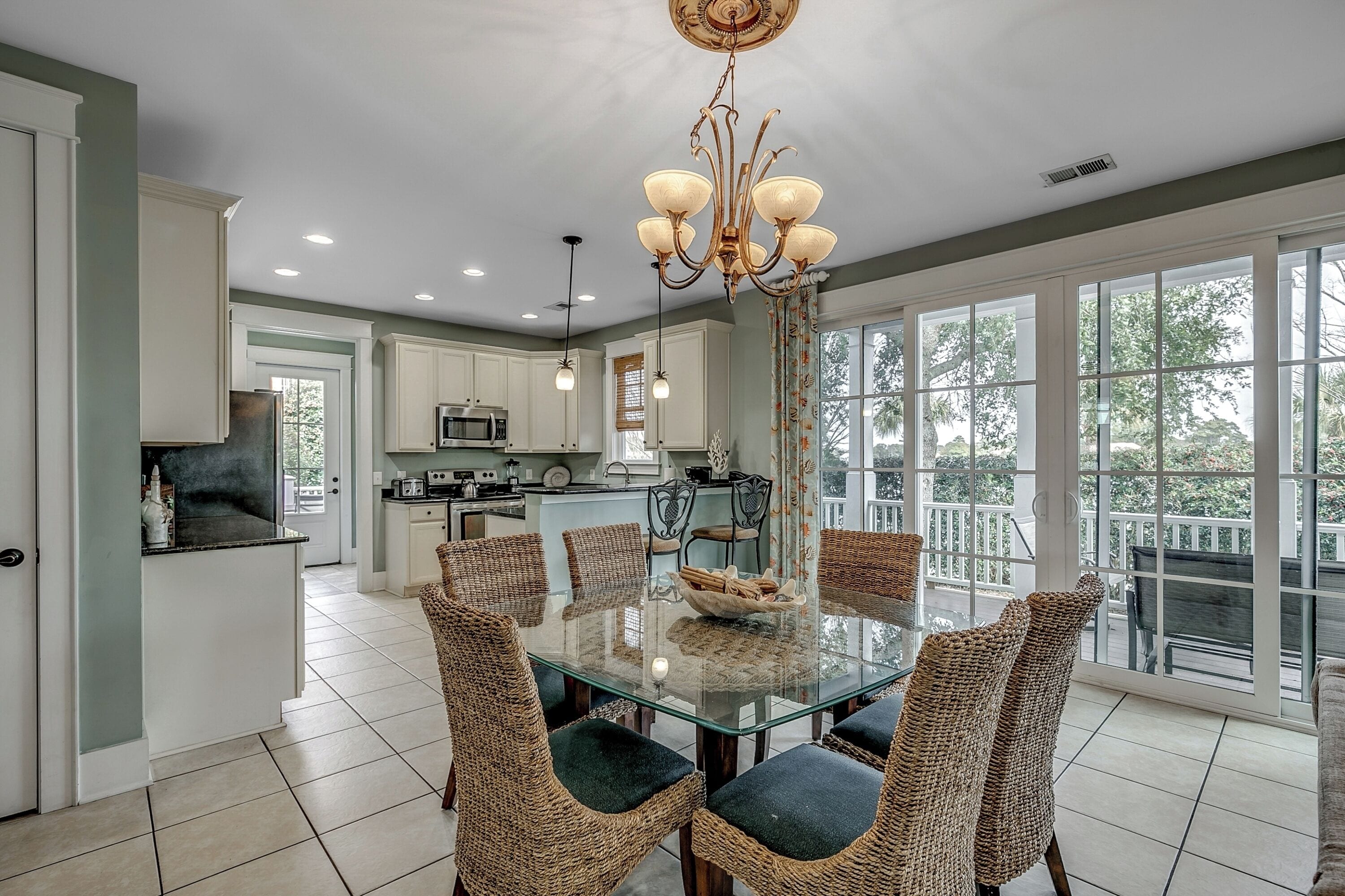 A spacious dining area with a glass table and wicker chairs, a kitchen in the background, and large windows offering a view of the outdoors.