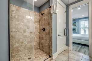 Un baño moderno con ducha acristalada, una puerta blanca y una habitación visible con cama y ventana.