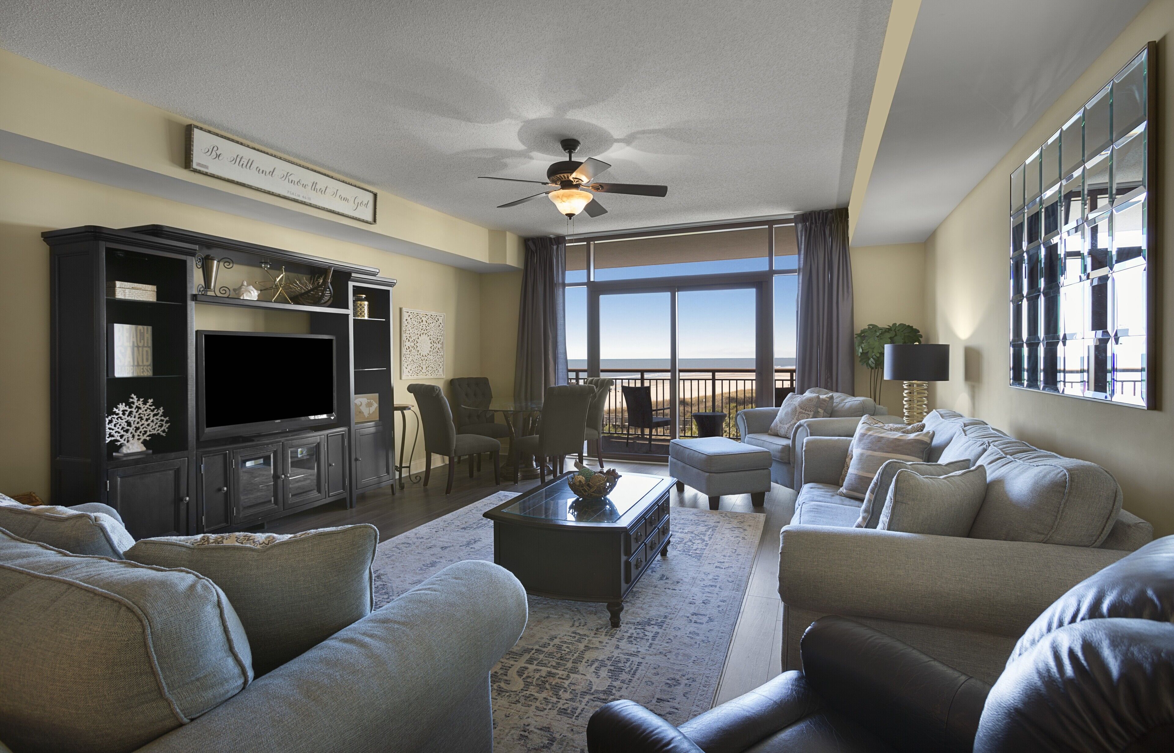 A spacious living room with a large flat-screen TV, comfortable seating, a ceiling fan, and a view of the outdoors.