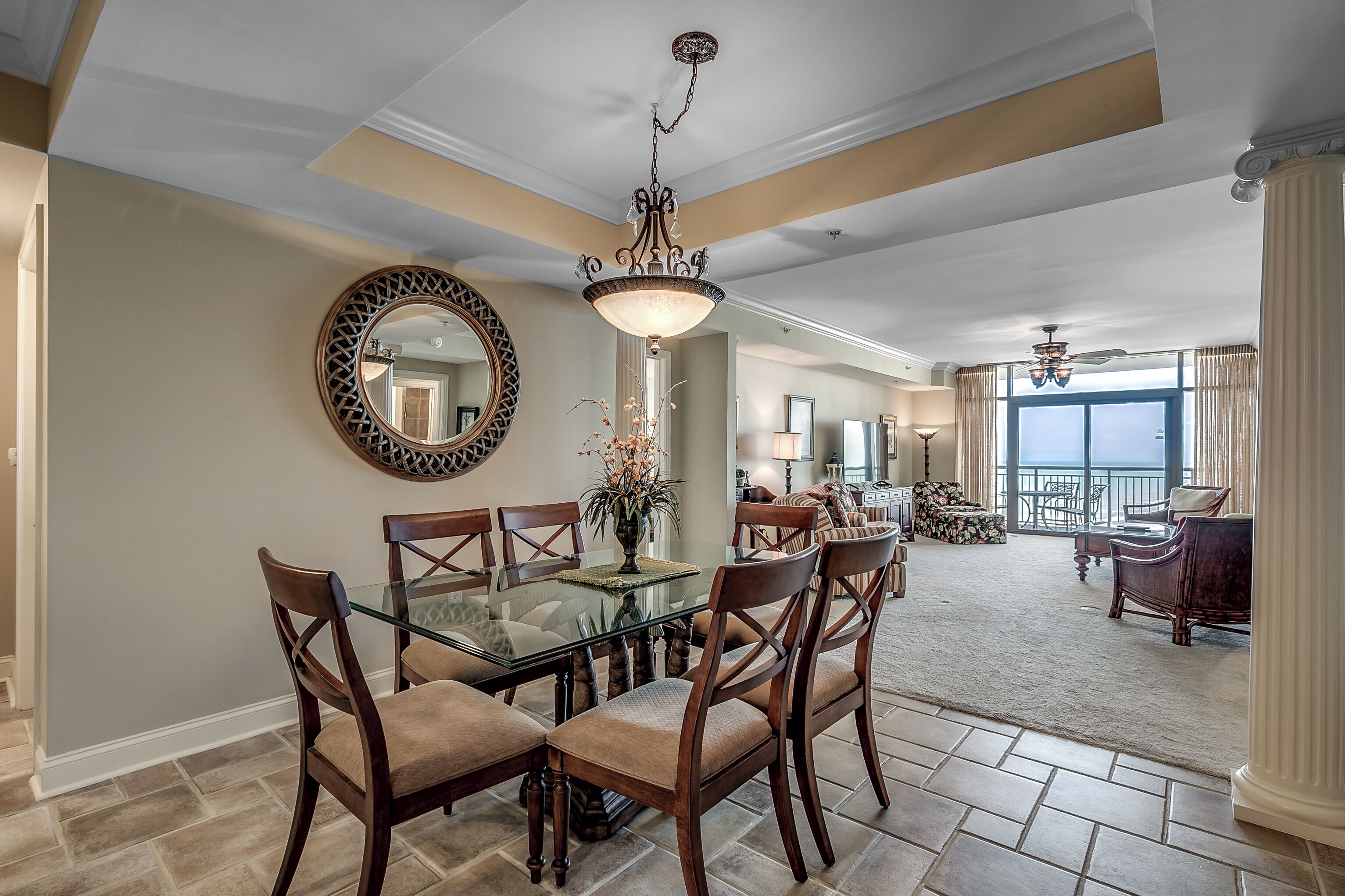 A dining area with a glass table and wooden chairs, a chandelier, and a large mirror on the wall.