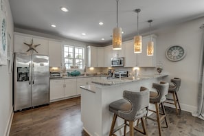 A modern kitchen with white cabinetry, stainless steel appliances, a central island with bar stools, pendant lighting, and a clock on the wall.