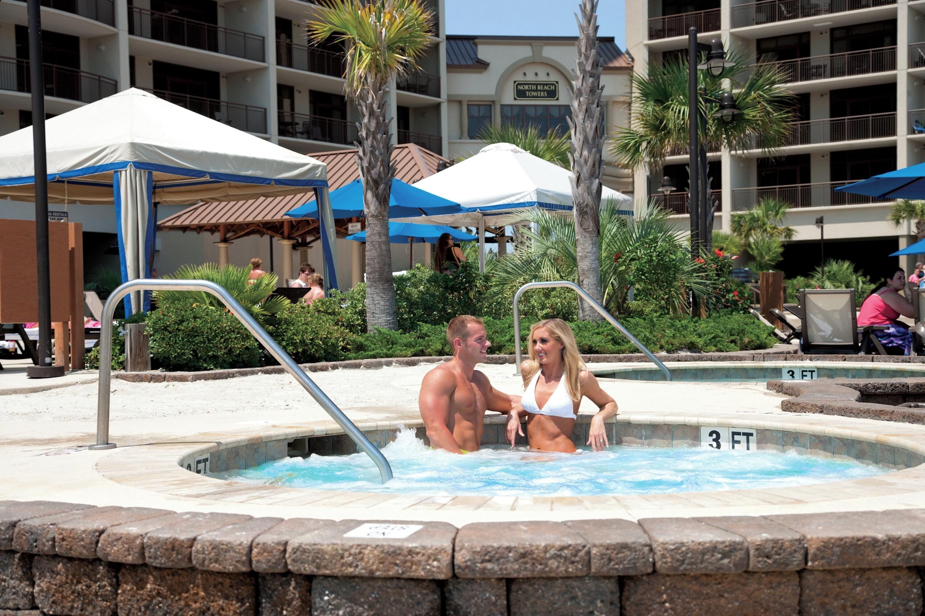A swimming pool area with a hot tub, palm trees, and a building labeled "North Beach Towers" in the background.