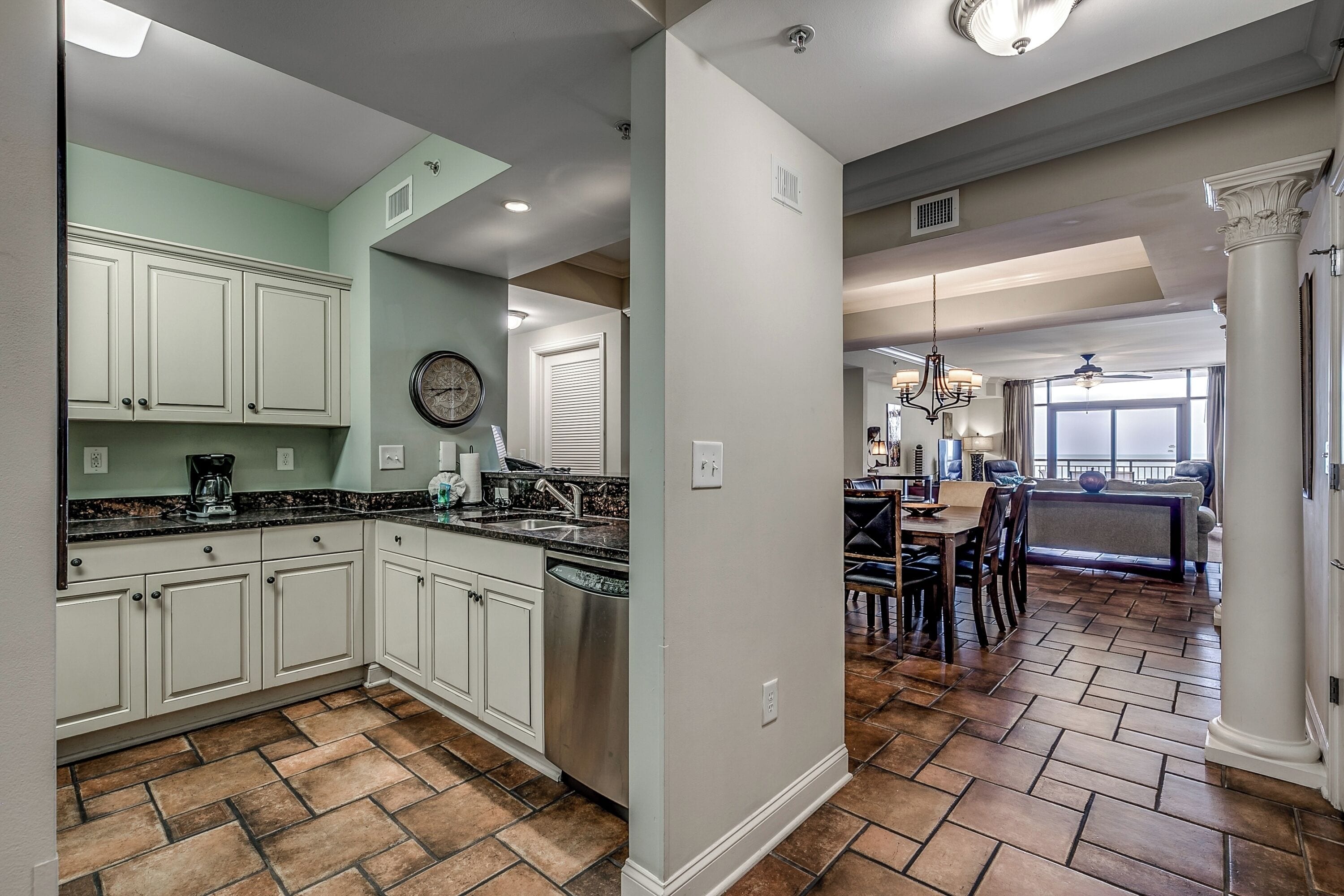 A kitchen with white cabinets, a black countertop, and a dishwasher. There is a dining area with a table and chairs, and a large window providing natural light.