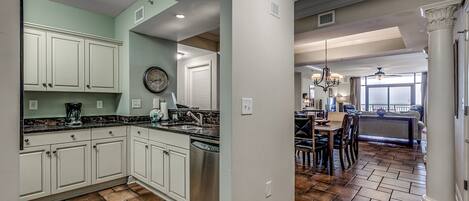 A kitchen with white cabinets, a black countertop, and a dishwasher. There is a dining area with a table and chairs, and a large window providing natural light.