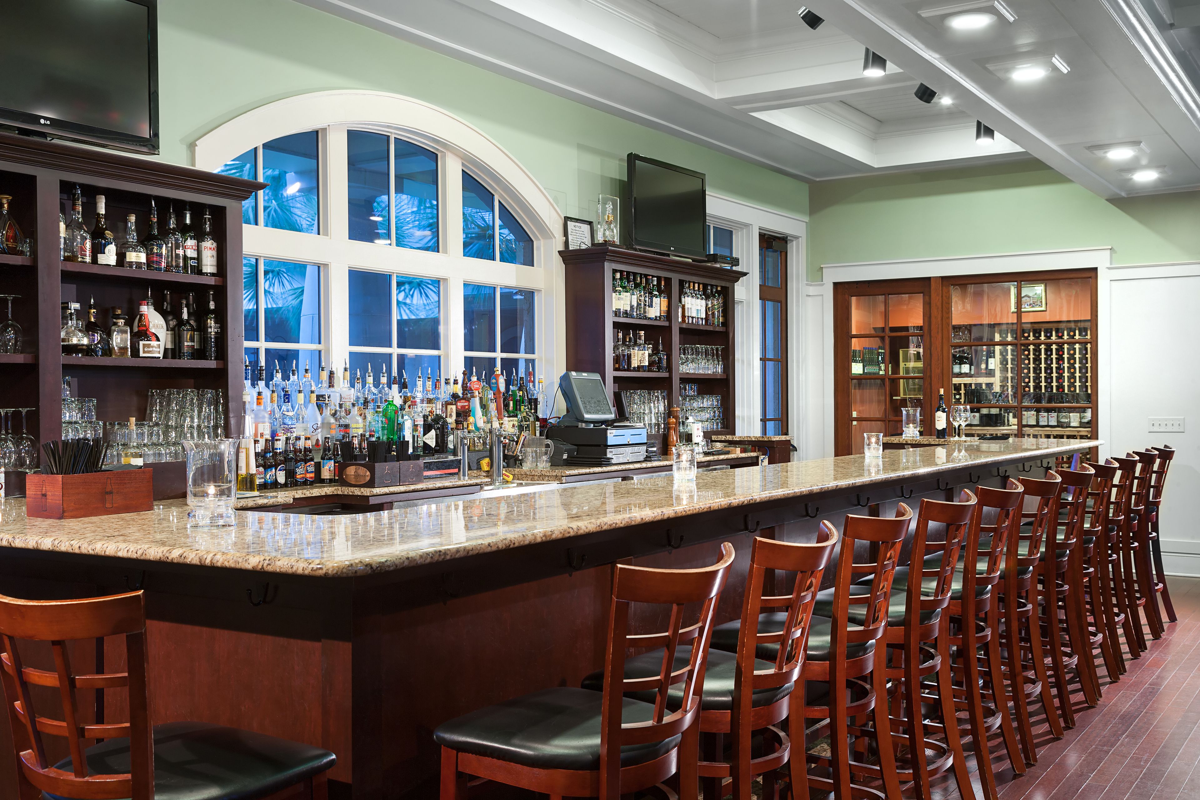 A bar with a long counter, multiple shelves of bottles, and a large window.