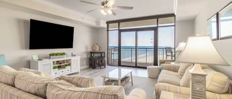 A spacious living room with a large flat-screen TV, striped sofas, a ceiling fan, and a balcony with a view of the ocean.