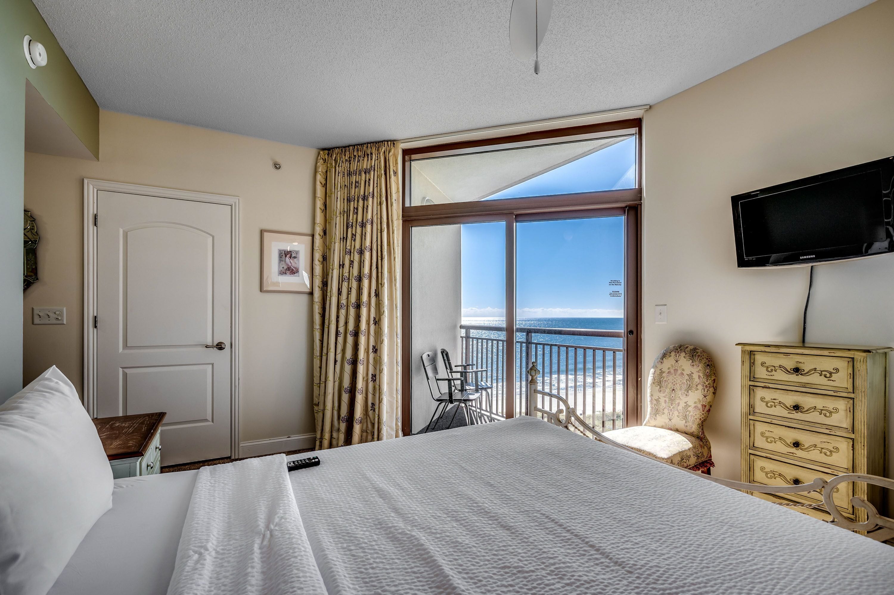 A hotel room with a bed, a dresser, a chair, a television, and a balcony with a view of the ocean.