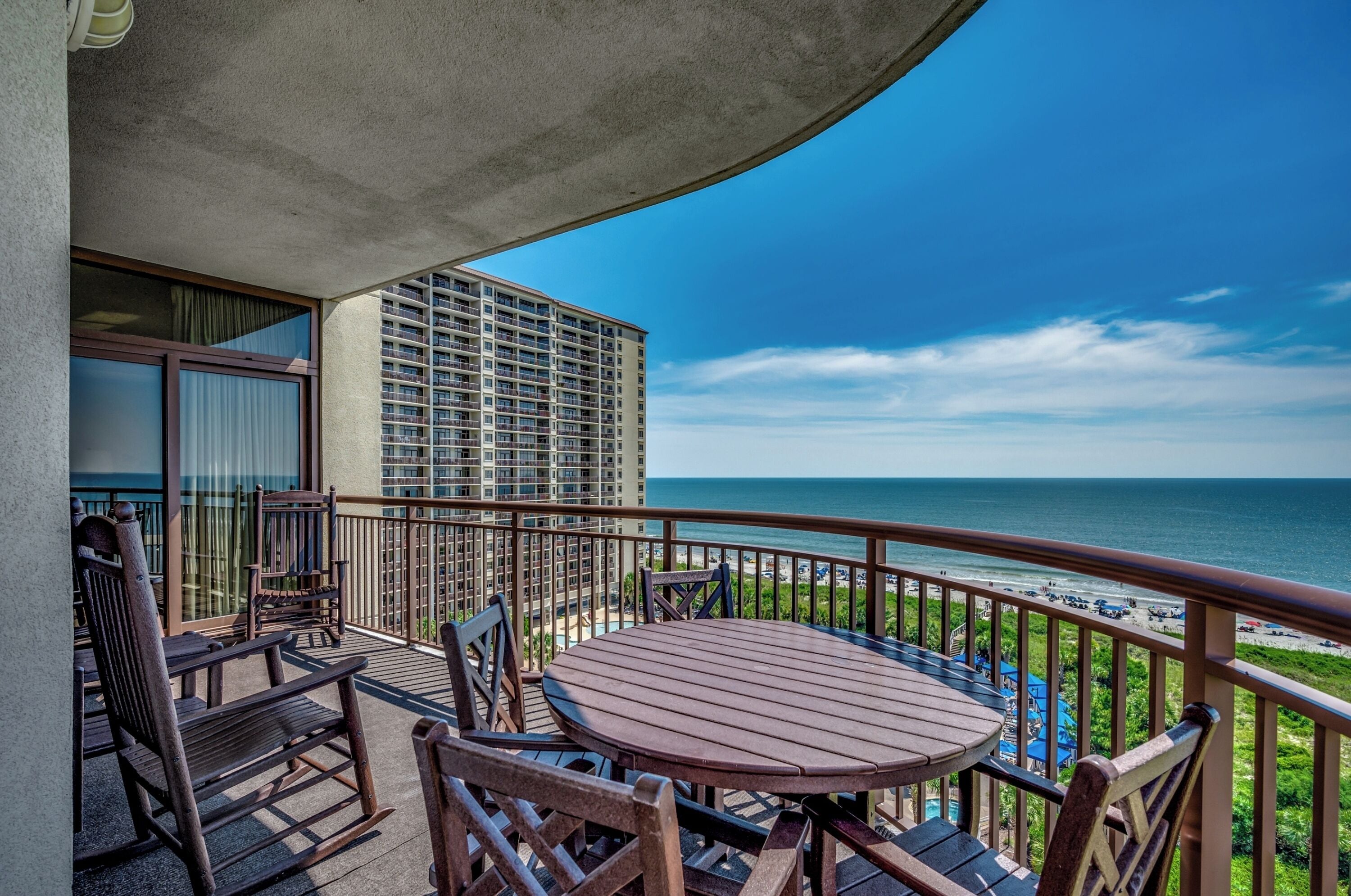 A balcony with a wooden table and chairs, overlooking a beach and a high-rise building.