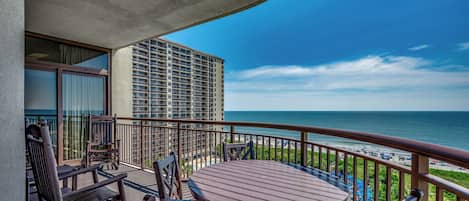 A balcony with a wooden table and chairs, overlooking a beach and a high-rise building.