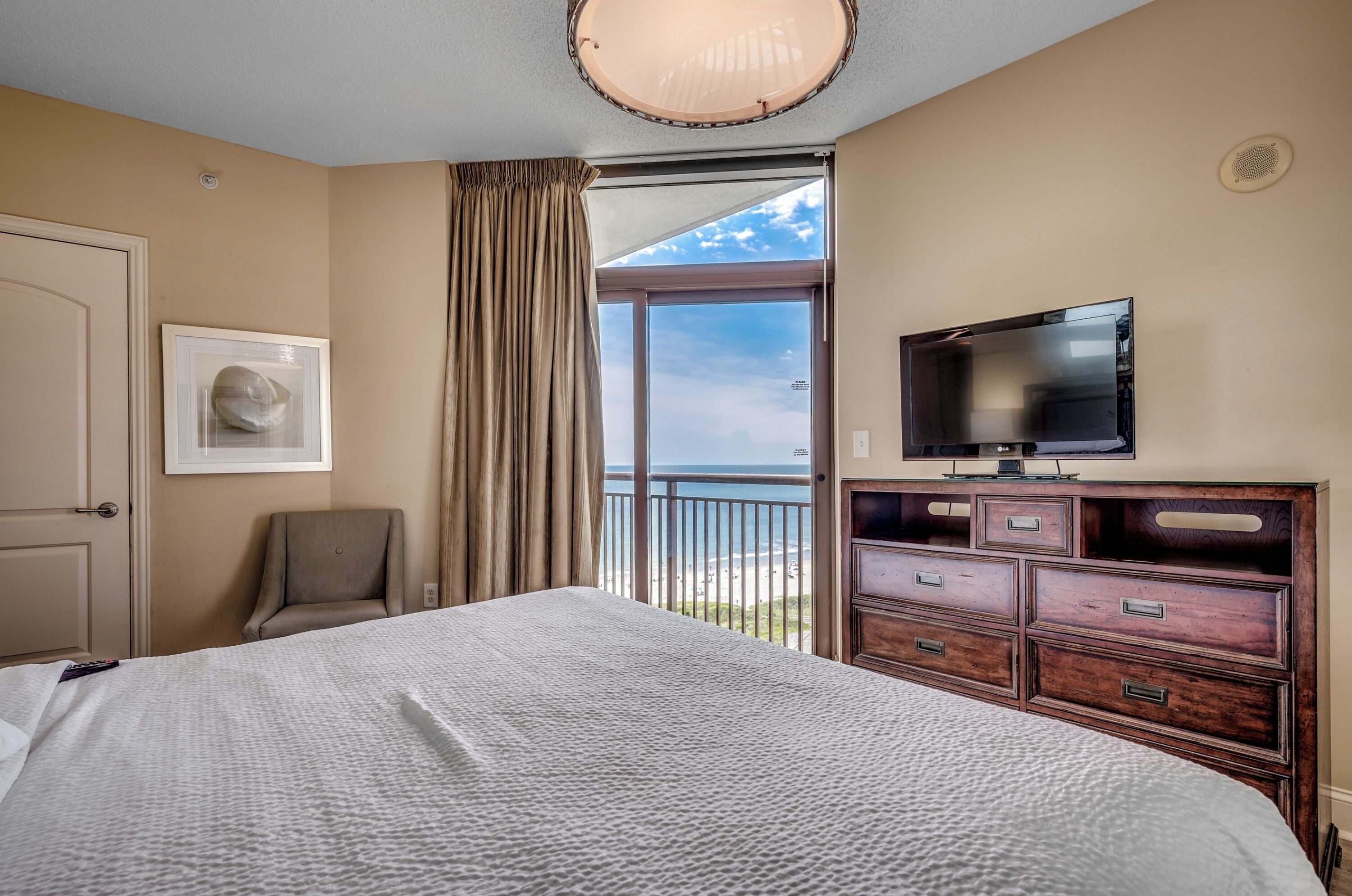A hotel room with a large bed, a TV, a chair, and a view of the beach.