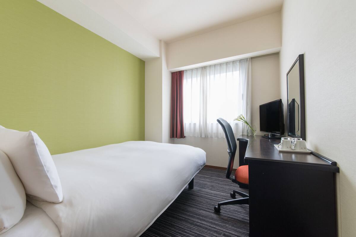 Une chambre d’hôtel avec un lit, un bureau avec un ordinateur, une chaise et une fenêtre avec des rideaux.