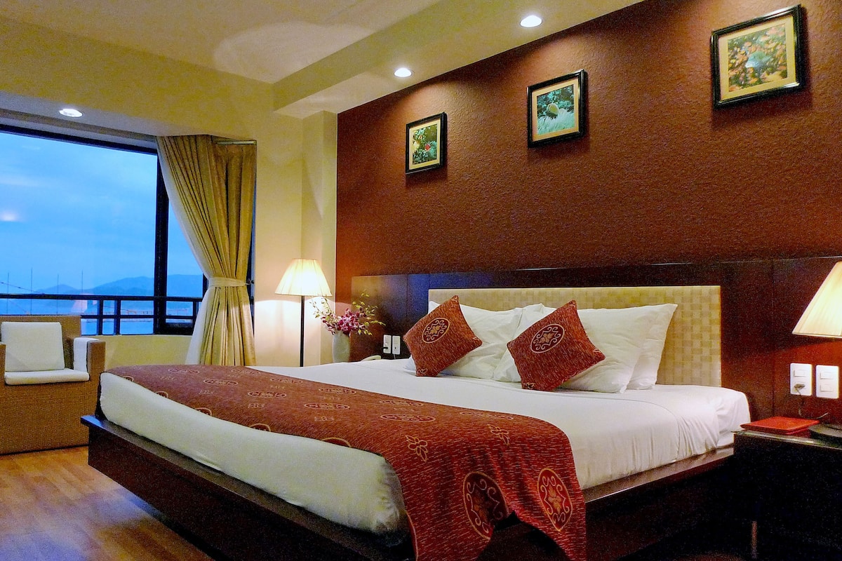 A hotel room with a large bed, a desk, a chair, and a view of the sea.