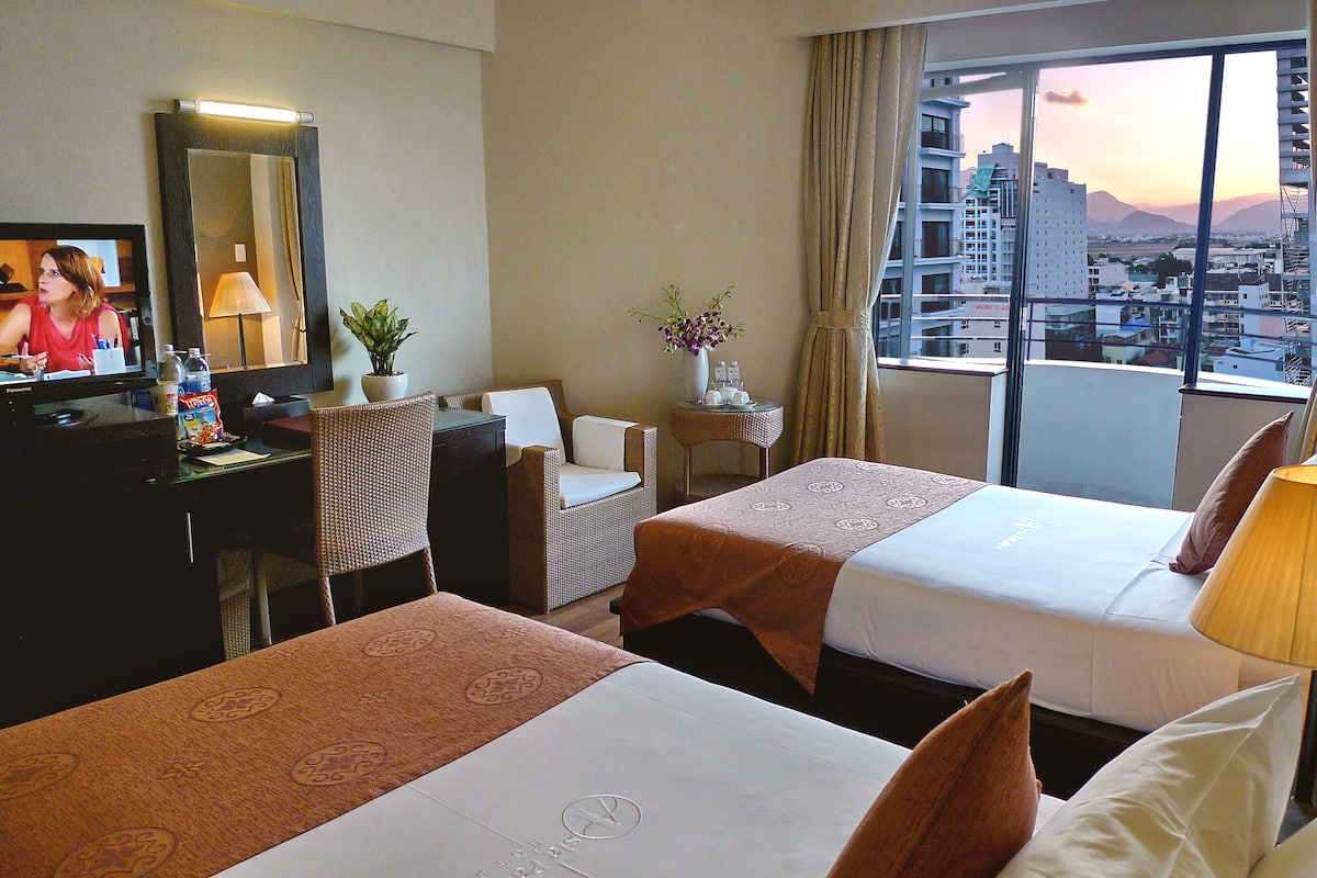 A hotel room with two beds, a desk with a computer, a chair, a TV, and a view of the cityscape.