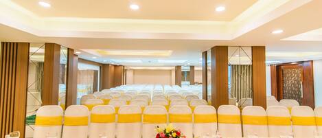 Un salón de conferencias con filas de sillas blancas con cintas amarillas, una mesa larga con vasos de agua y un centro de mesa floral.
