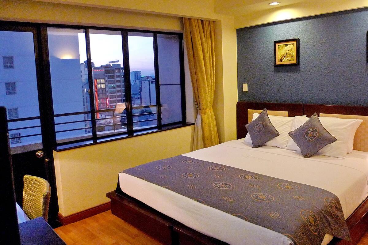 A hotel room with a large bed, two pillows, a nightstand with a lamp, a window with curtains, and two framed pictures on the wall.