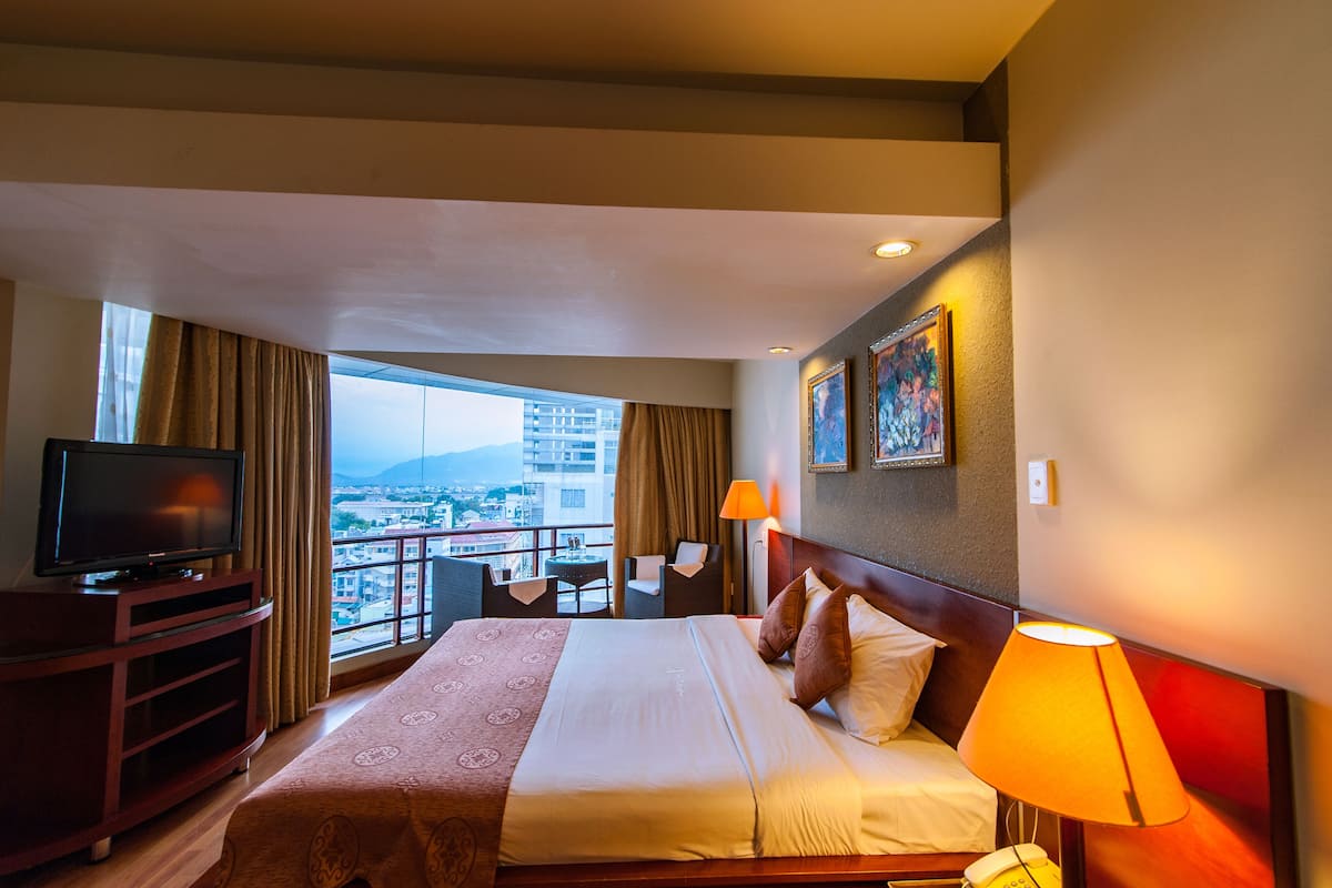 A hotel room with a large bed, a TV, a balcony with a view of the city, and a painting on the wall.