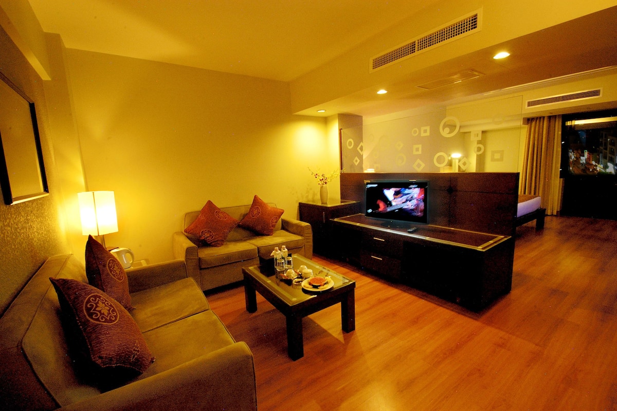 A hotel room with a sofa, a coffee table, a TV, and a minibar.
