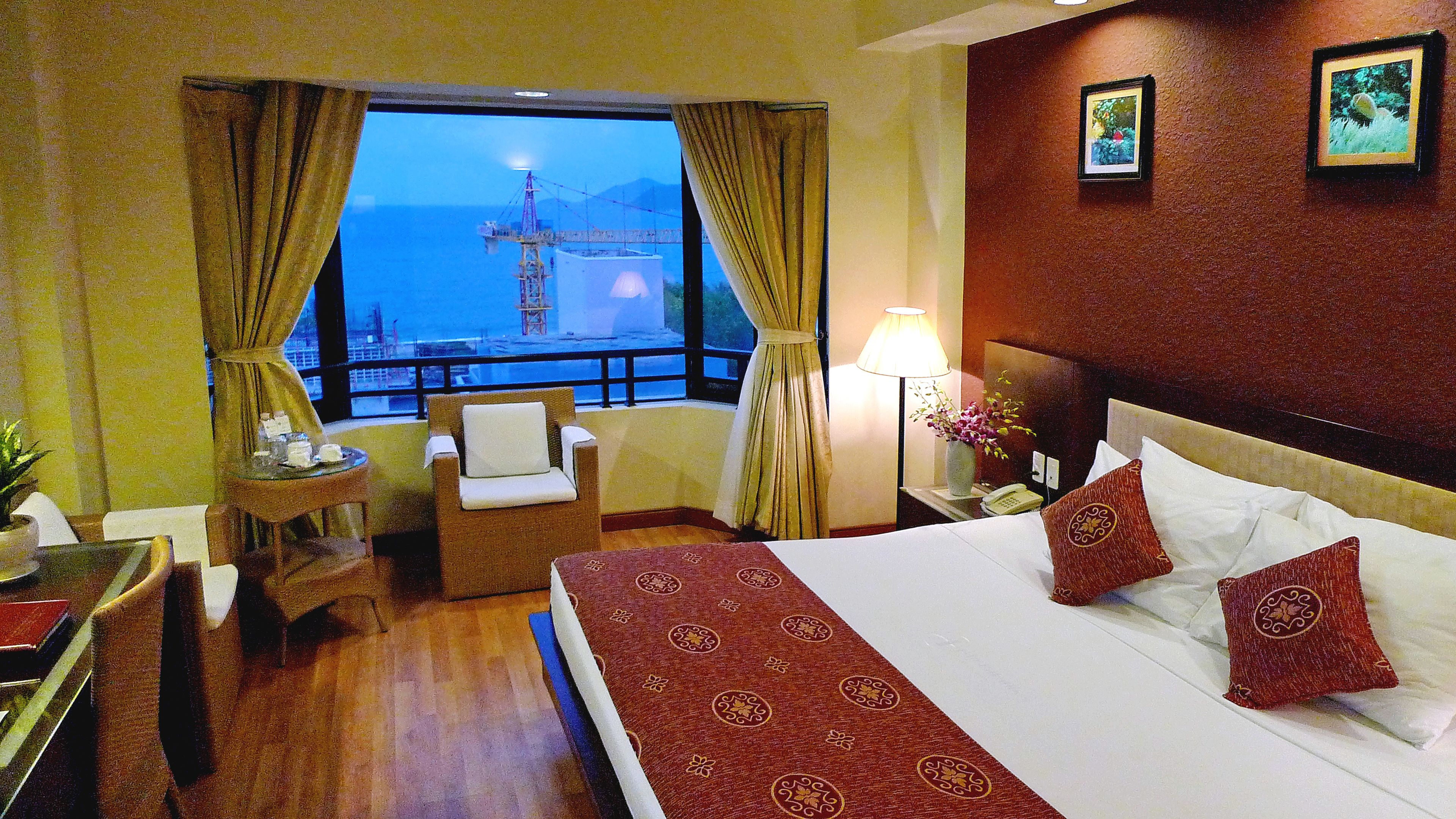 A hotel room with a large bed, a chair, a small table, a lamp, and a view of the sea with a crane.