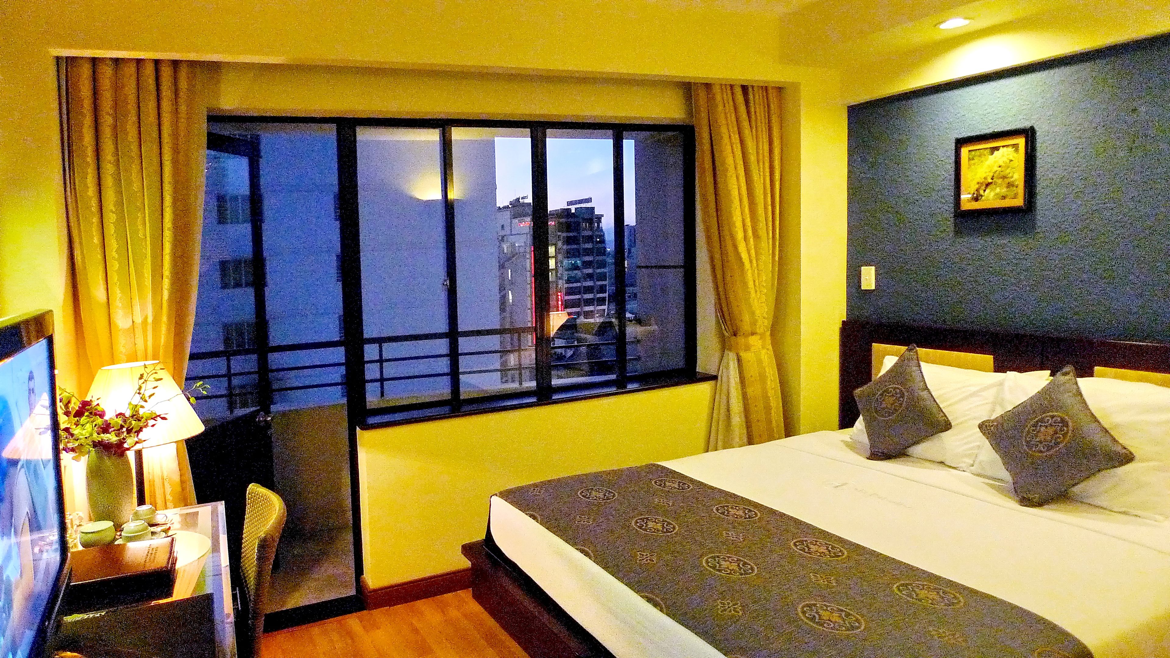 Una habitación de hotel con un ventanal grande, una cama con ropa de cama estampada, una mesita de noche con una lámpara y un cuadro en la pared.