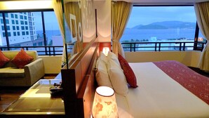 A hotel room with a bed, a sofa, a table, and a view of the sea and buildings. - Asia Paradise Hotel (Nha Trang)