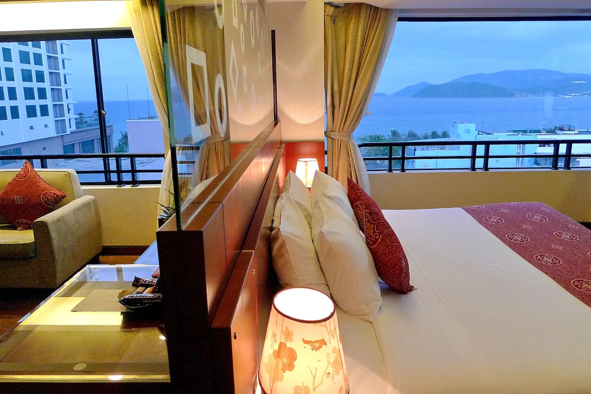 A hotel room with a bed, a sofa, a table, and a view of the sea and buildings.