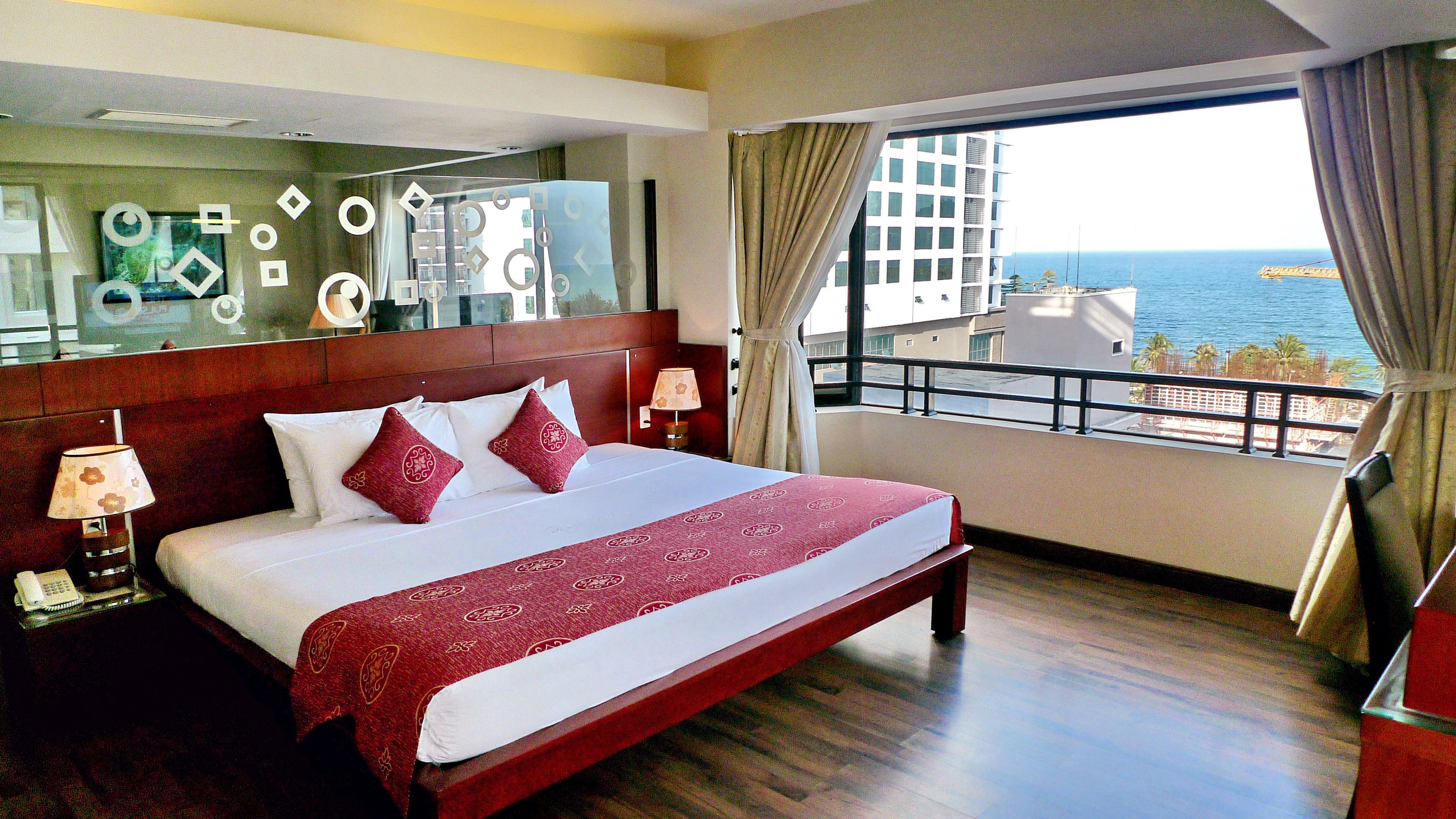 A hotel room with a large bed, a nightstand, a lamp, and a view of the ocean.