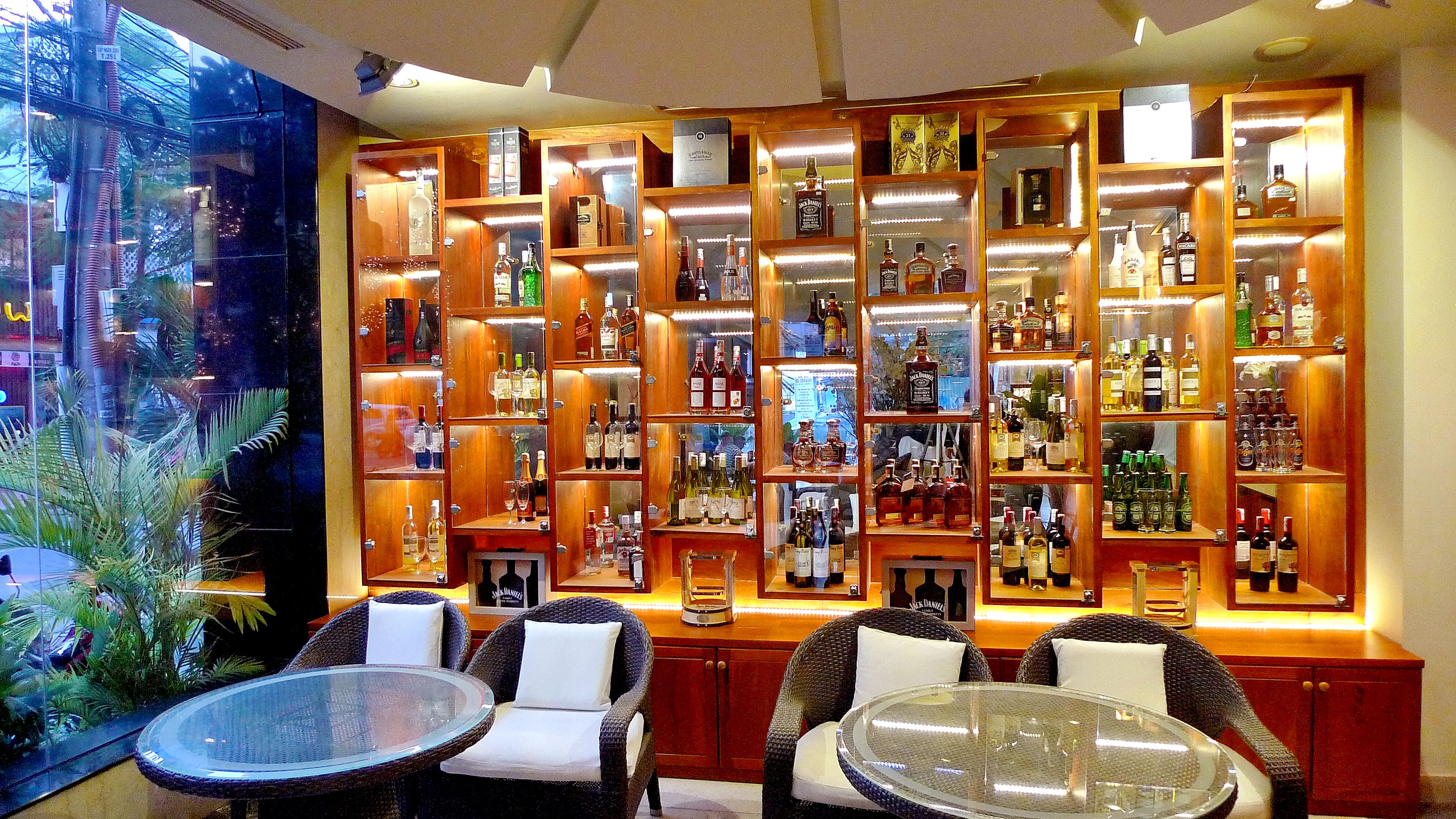 Una zona de bar con varias estanterías llenas de botellas de licores, sillas de mimbre y una mesa de vidrio.