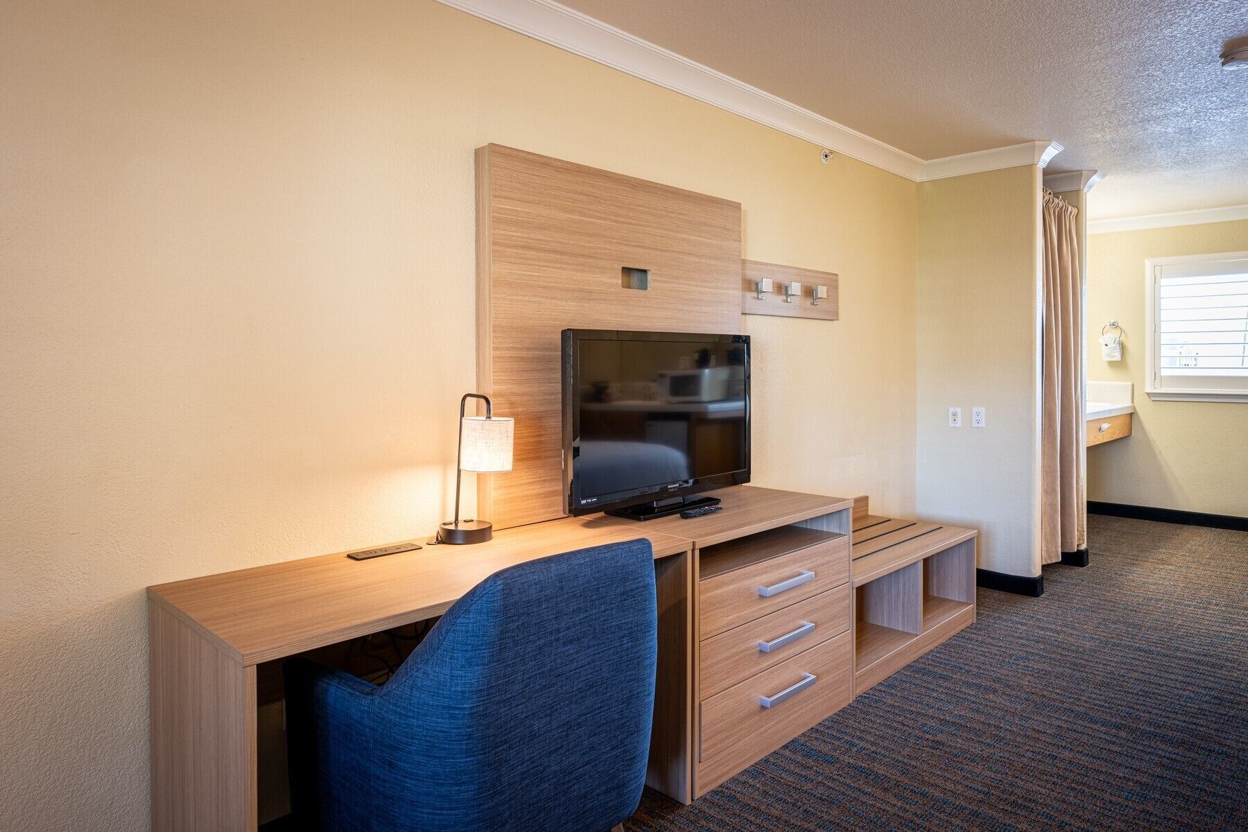 A hotel room with a wooden desk, a television, a lamp, a chair, and a wardrobe.