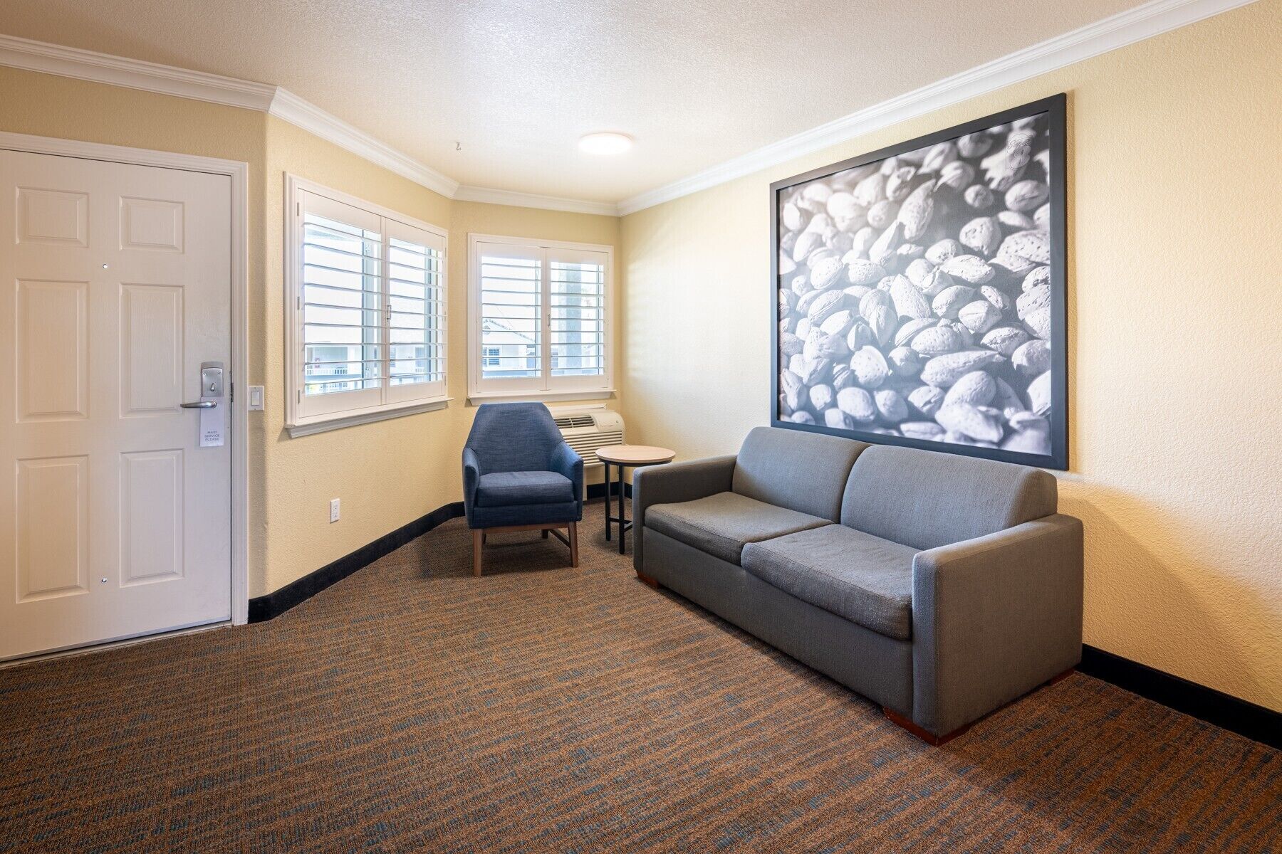Ein Hotelzimmer mit grauem Sofa, einem kleinen runden Tisch, einem blauen Stuhl und einem großen abstrakten Gemälde an der Wand.