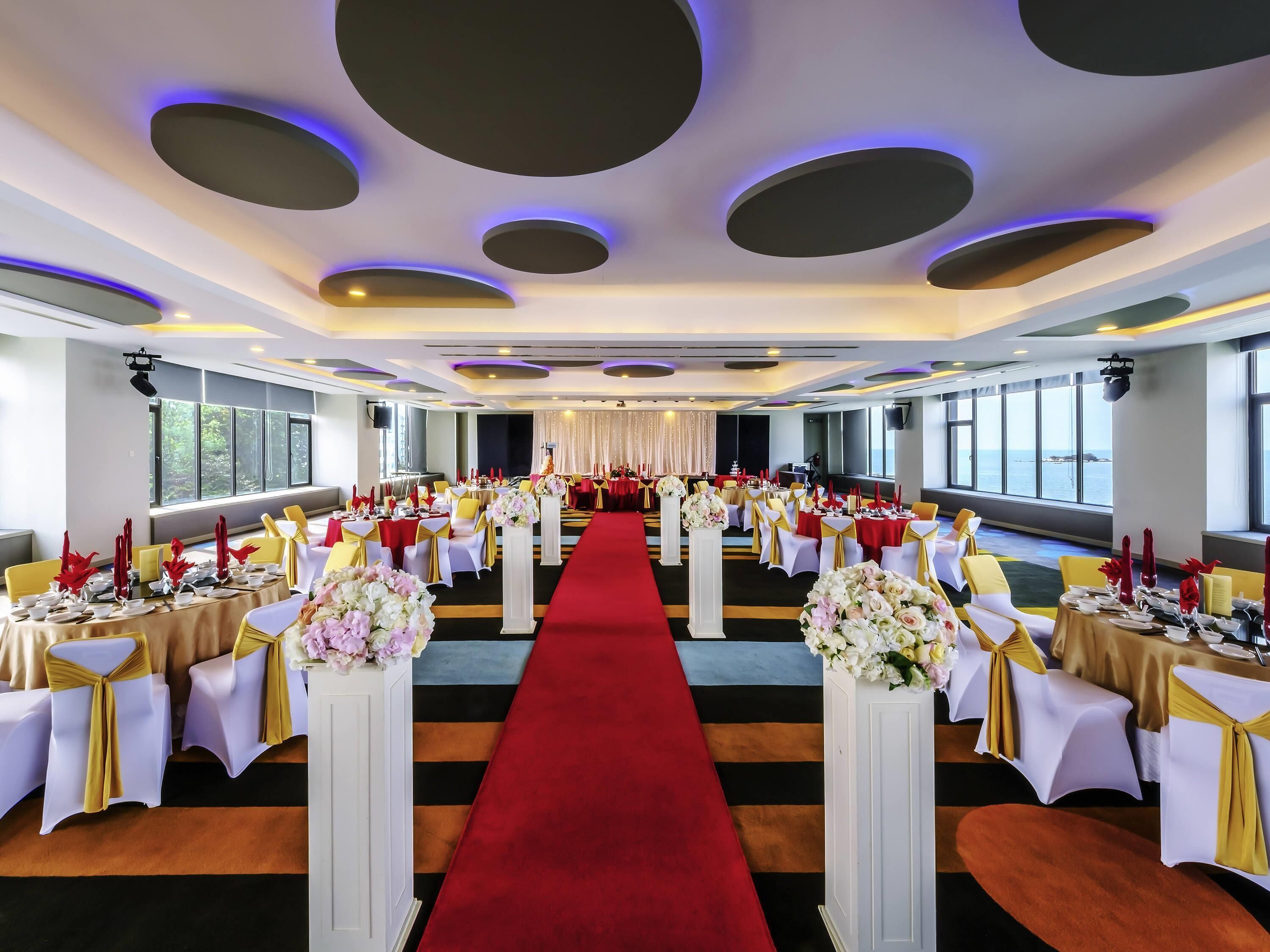 A banquet hall with tables set for a formal event, a red carpet, and a view of the sea through large windows.