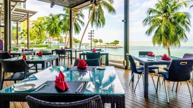 An outdoor dining area with tables and chairs, overlooking a beach and palm trees. - Mercure Penang Beach (George Town)