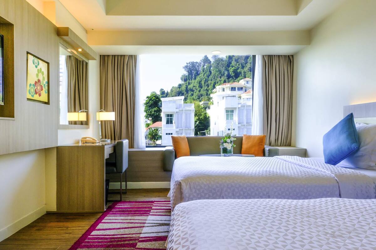 A hotel room with two beds, a desk, a chair, and a view of buildings and trees.