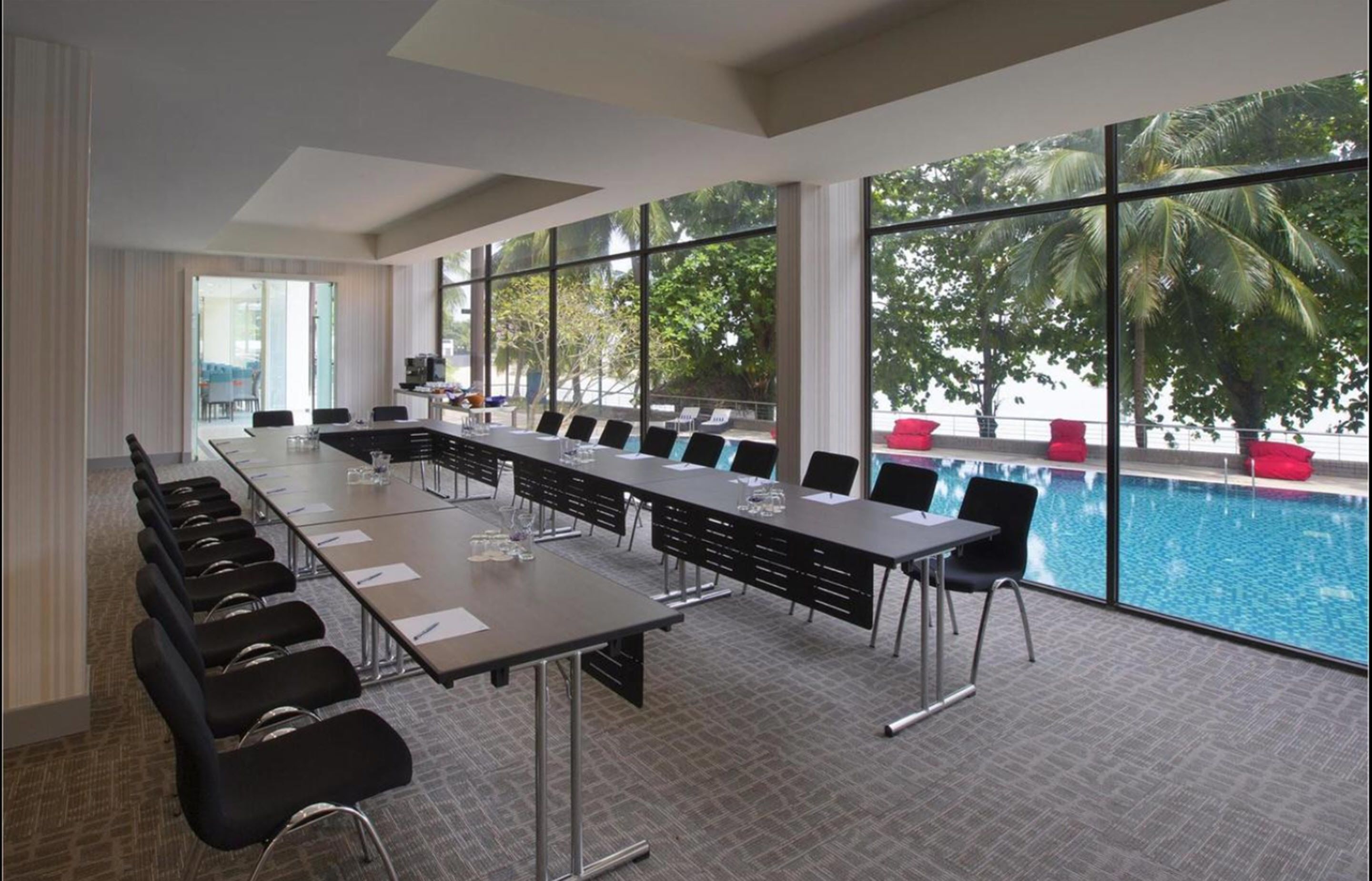 A conference room with a long table and chairs, large windows with a view of a pool and palm trees, and a carpeted floor.