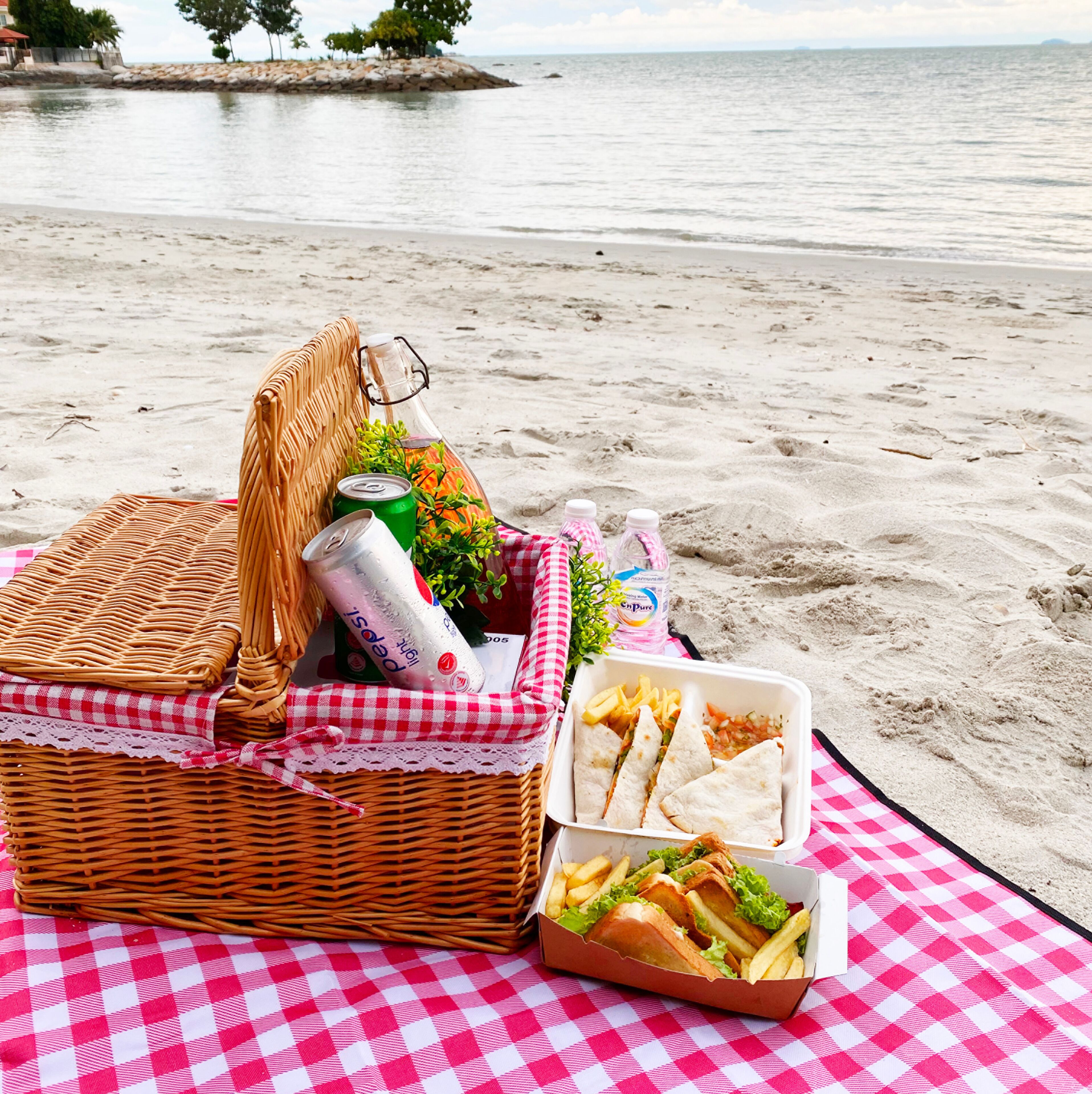 A picnic setup on a beach with a wicker basket containing a can, a bottle, and a plant.