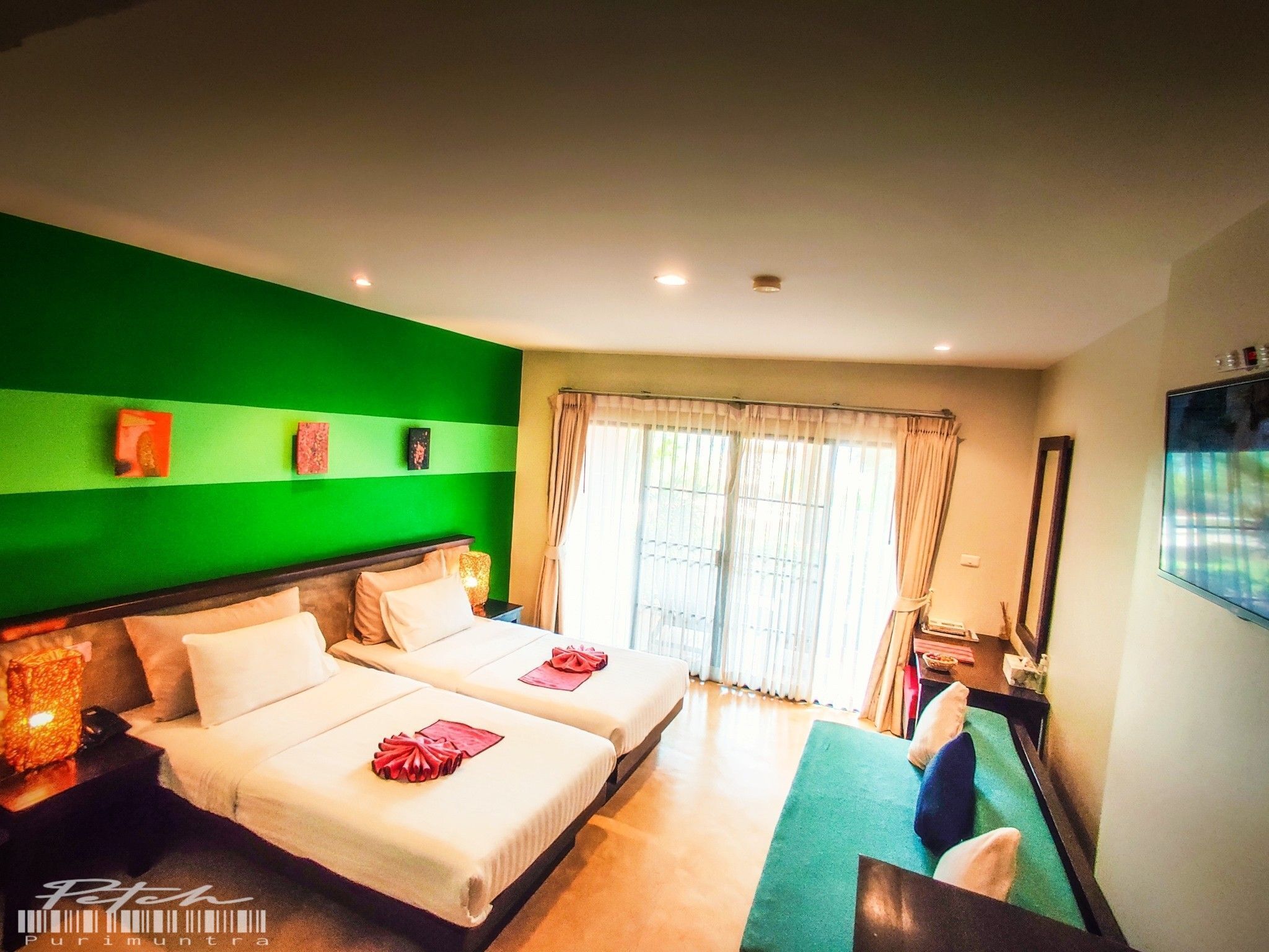 Habitación de hotel con dos camas, una pared decorada en verde, un ventanal con cortinas y un televisor de pantalla plana montado en la pared.