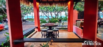 Un área de estar al aire libre con una mesa redonda y cuatro taburetes, rodeada de pilares rojos y plantas tropicales, con vista a un cuerpo de agua.