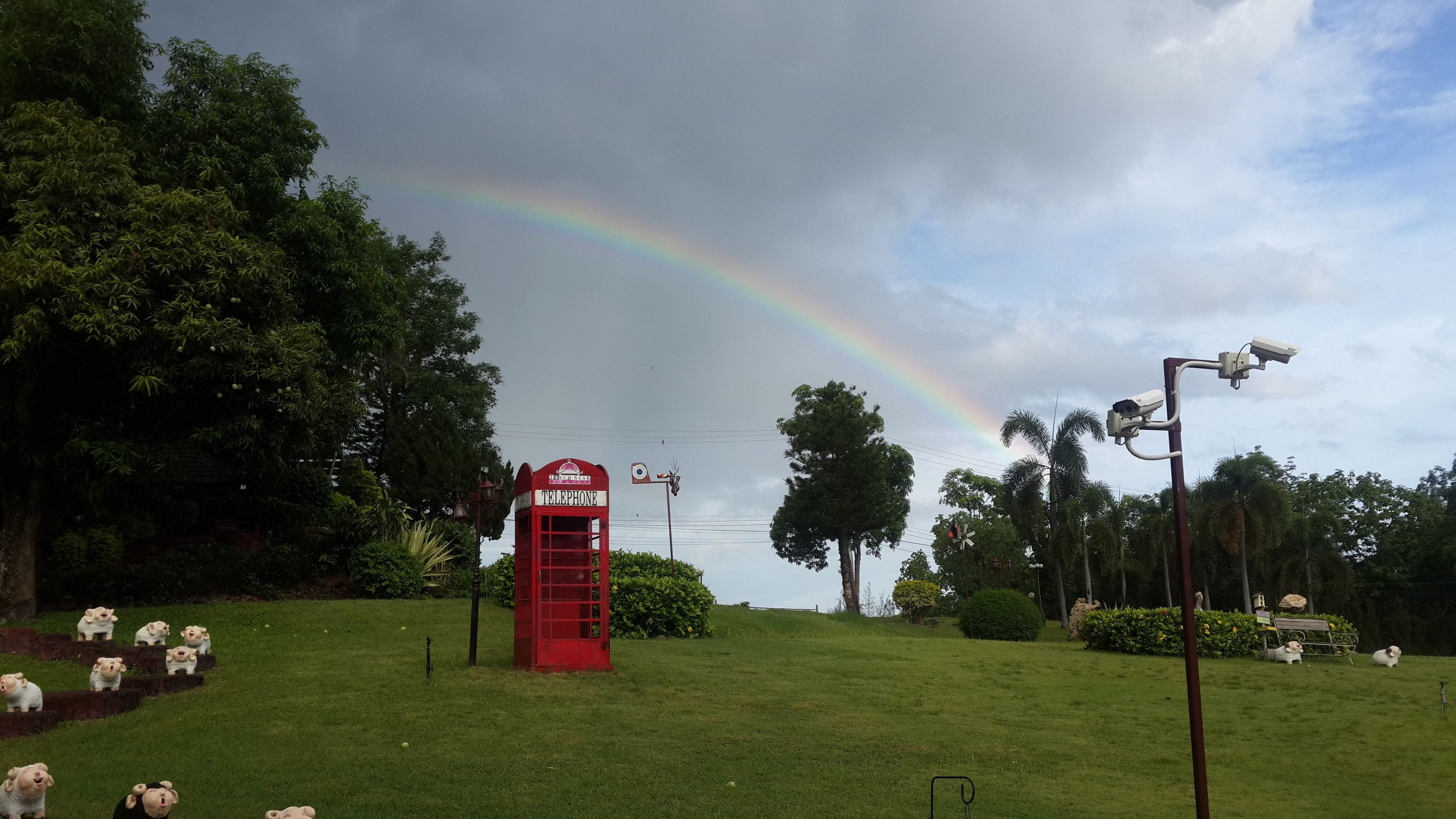 A red telephone booth in a grassy area with trees and a rainbow in the background.