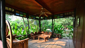 Una terraza de madera con ventilador de techo, mobiliario de madera y vista a exuberante vegetación.