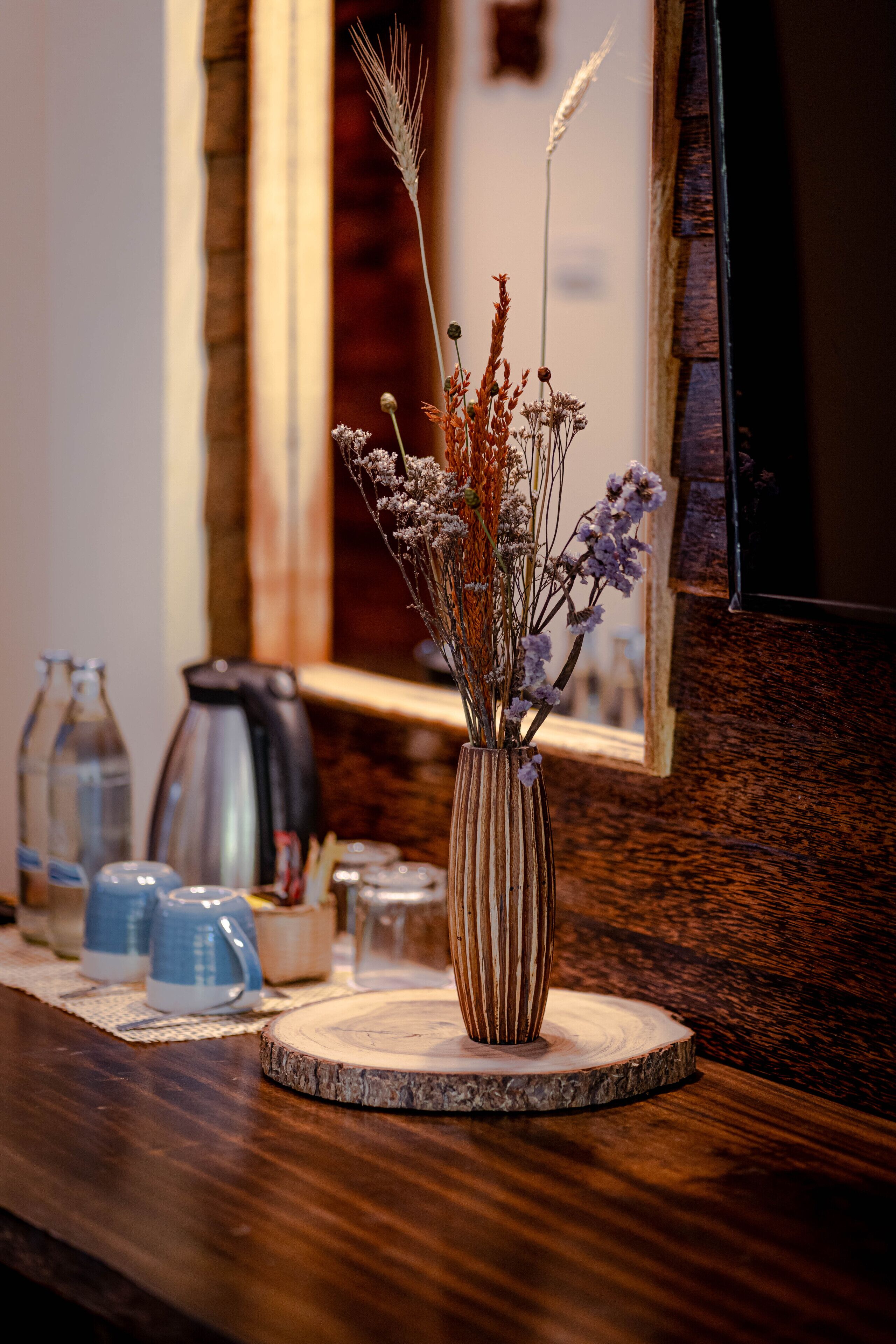 Une table en bois sur laquelle se trouvent un vase rempli de fleurs séchées, une bouilloire et divers récipients en verre.