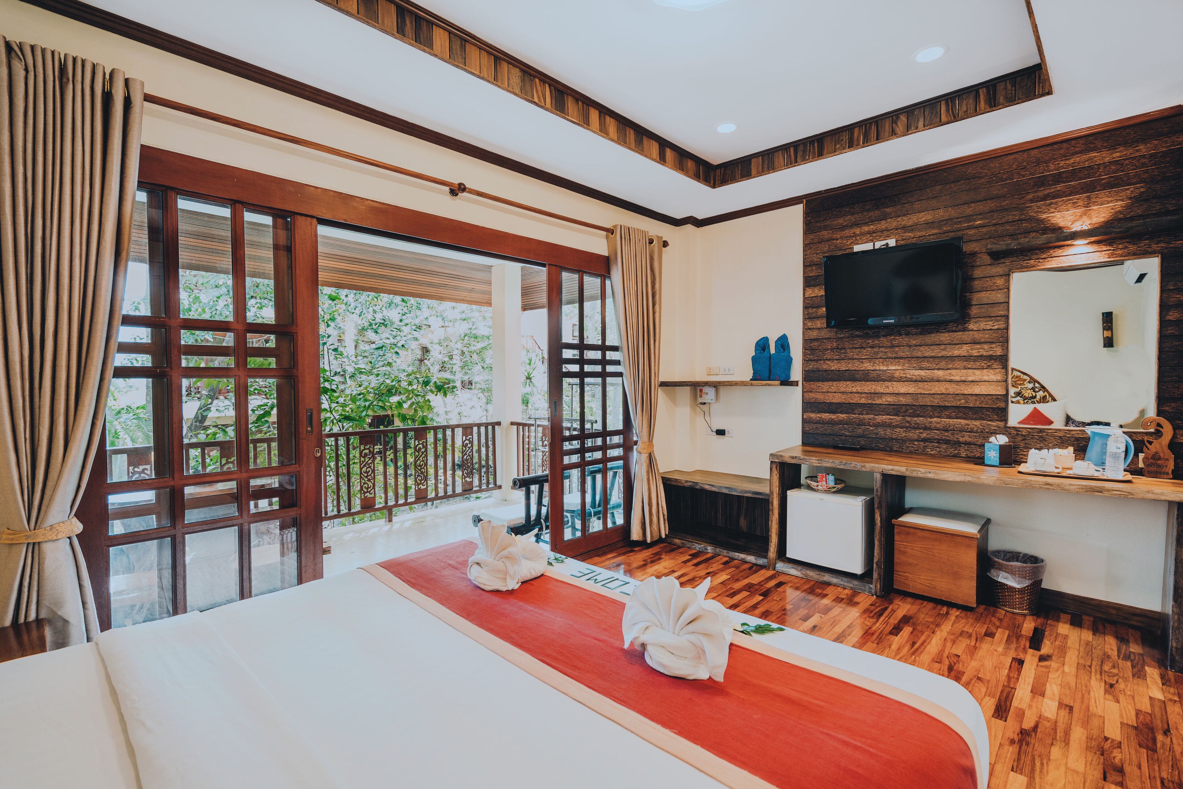 A hotel room with a large bed, wooden flooring, a desk with a TV, and a balcony with curtains.