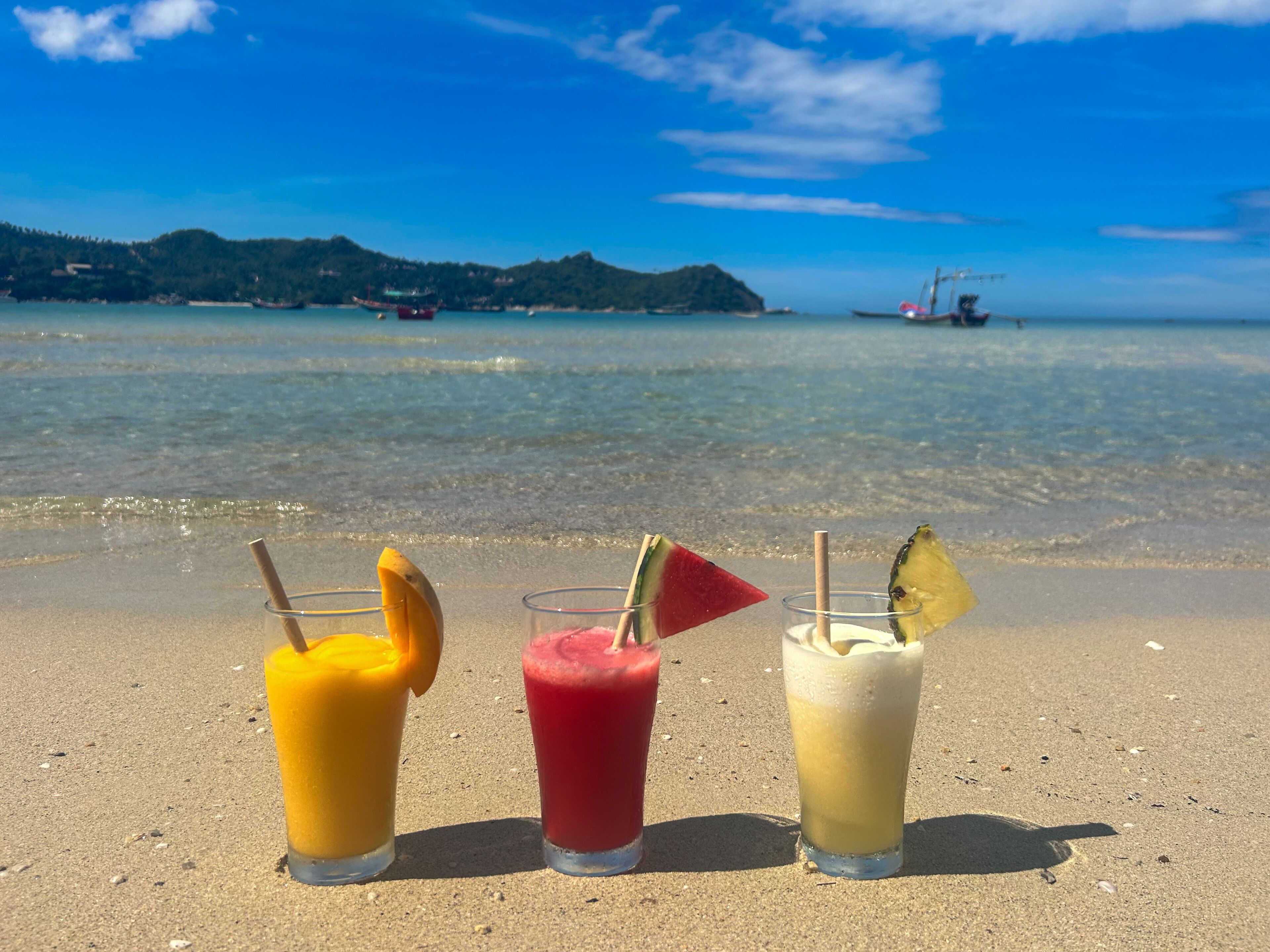 Tre bevande su una spiaggia sabbiosa con vista libera sull'oceano e barche in lontananza.