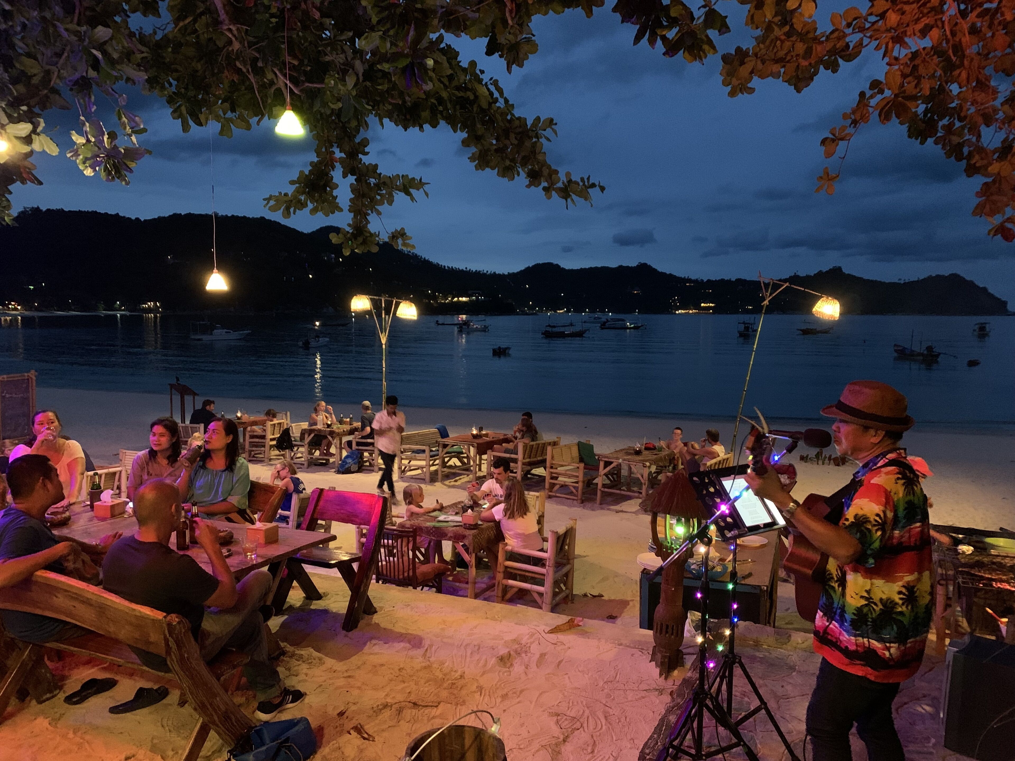 A beachside dining area with tables and chairs, a musician performing, and a view of the sea and boats in the background.