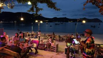 A beachside dining area with tables and chairs, a musician performing, and a view of the sea and boats in the background.