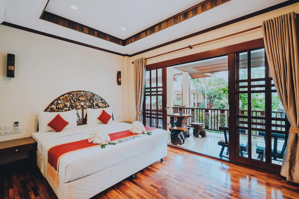 A hotel room with a large bed, wooden flooring, a balcony with a table and chairs, and a view of greenery outside.
