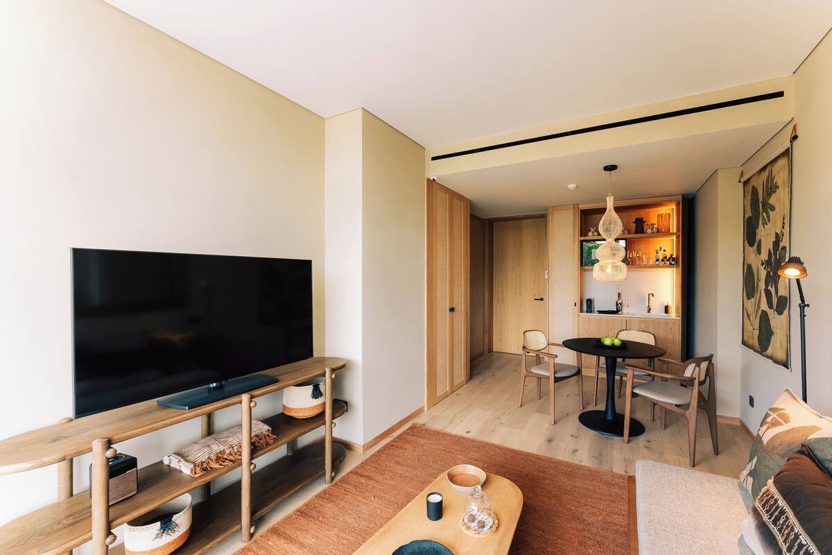 A modern living room with a flat-screen TV, wooden furniture, a dining area with a table and chairs, and a kitchen area with wooden cabinets and a hanging light fixture.