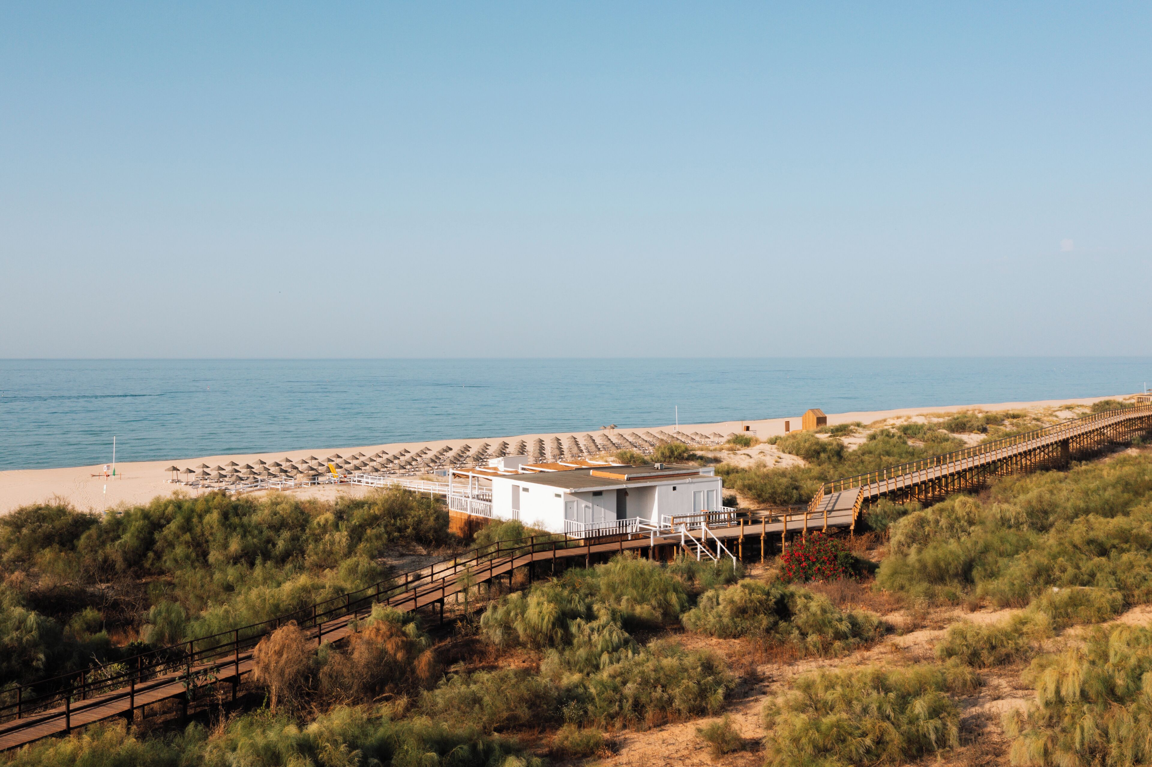 Un hotel frente a la playa con un edificio blanco, una terraza de madera y una playa arenosa con sombrillas.