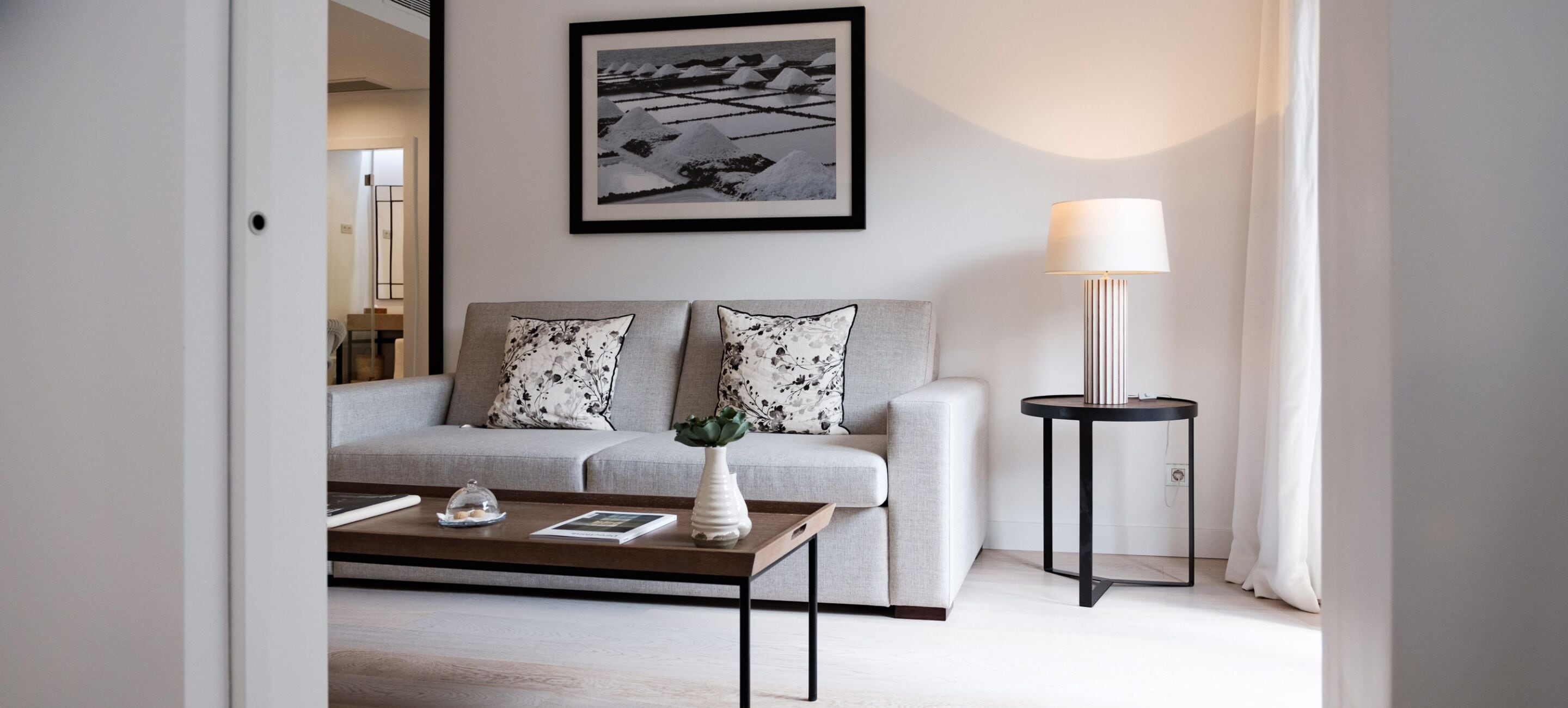 A modern living room with a grey sofa, two floral cushions, a coffee table with a vase and remote control, a framed picture on the wall, and a floor lamp.