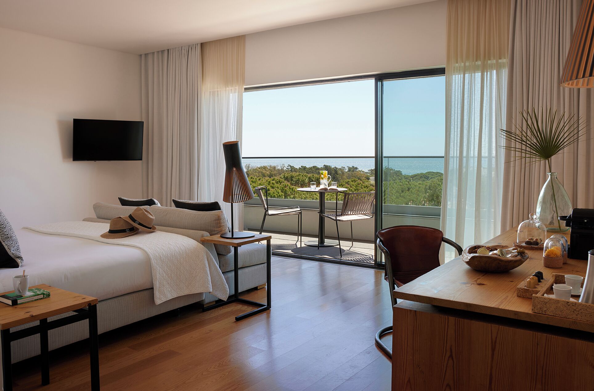 A hotel room with a bed, a desk, a chair, a table, a lamp, and a view of the ocean.