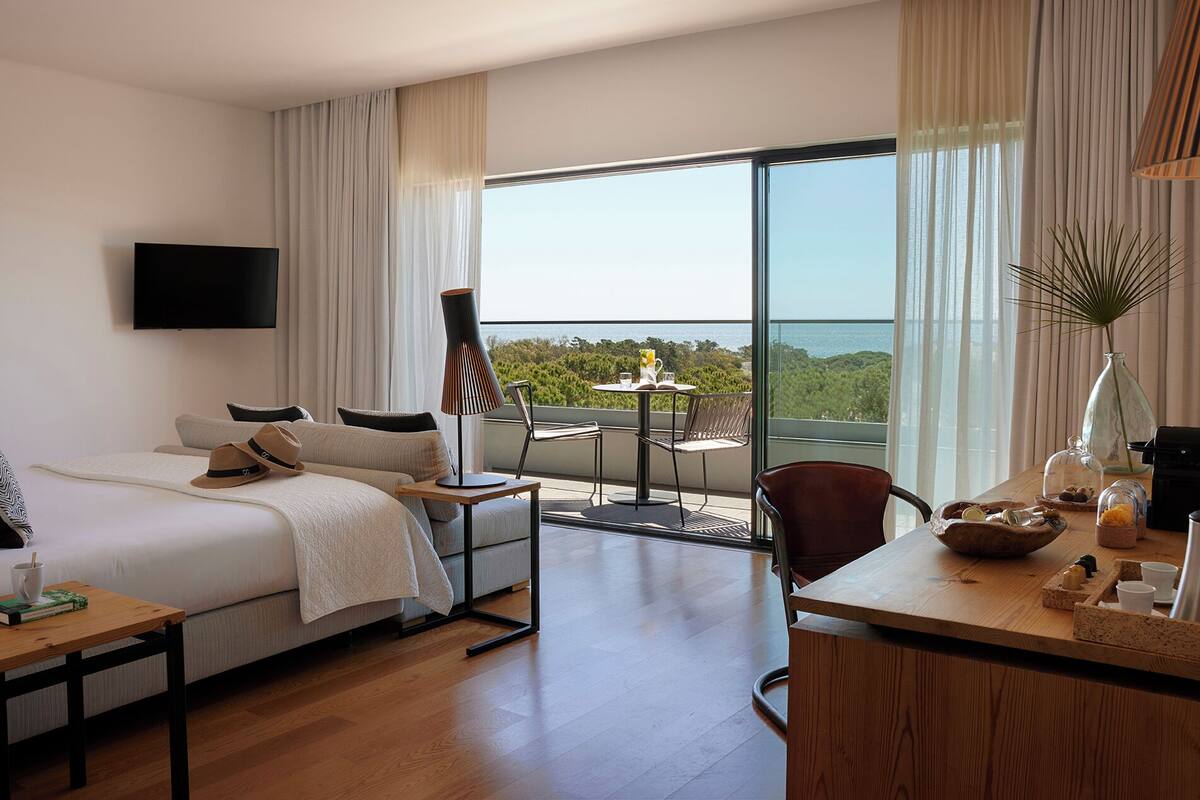A hotel room with a bed, a desk, a chair, a table, a lamp, and a view of the ocean.