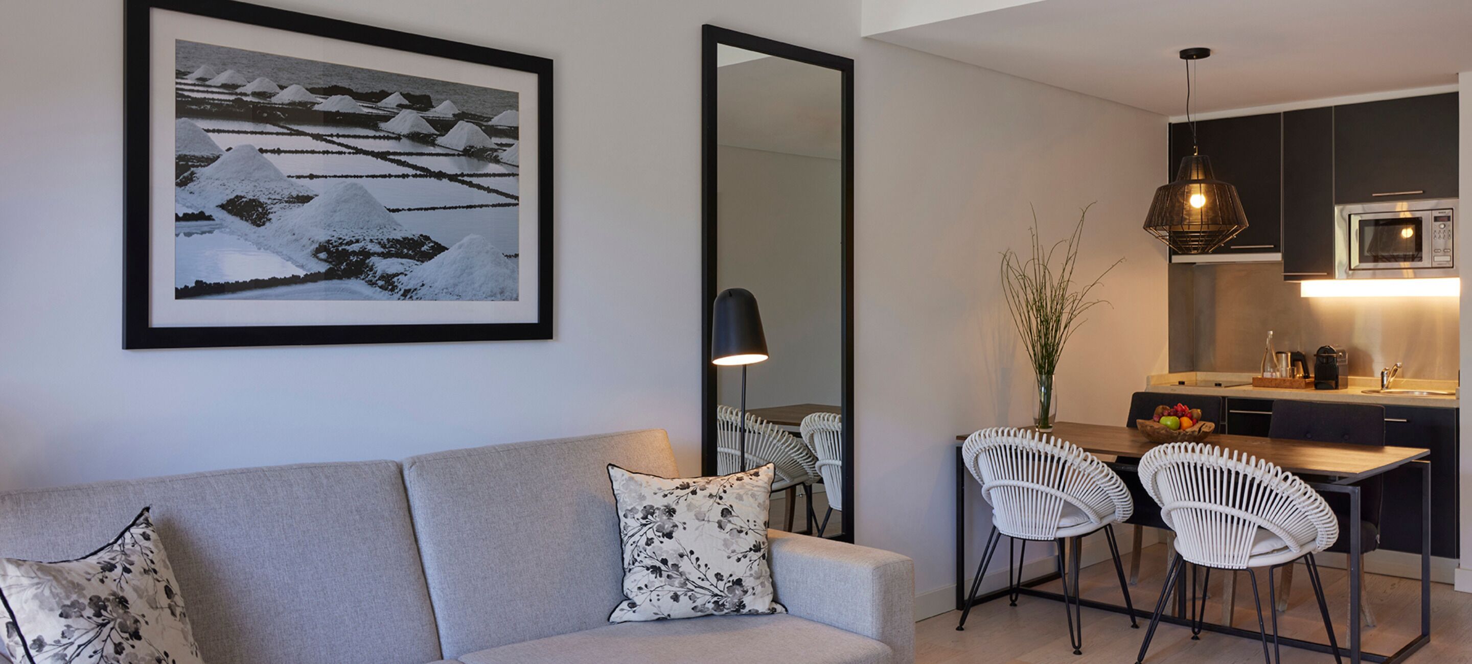 A modern living room with a sofa, two white chairs, a dining table, a kitchen area with a microwave, and a framed photograph on the wall.